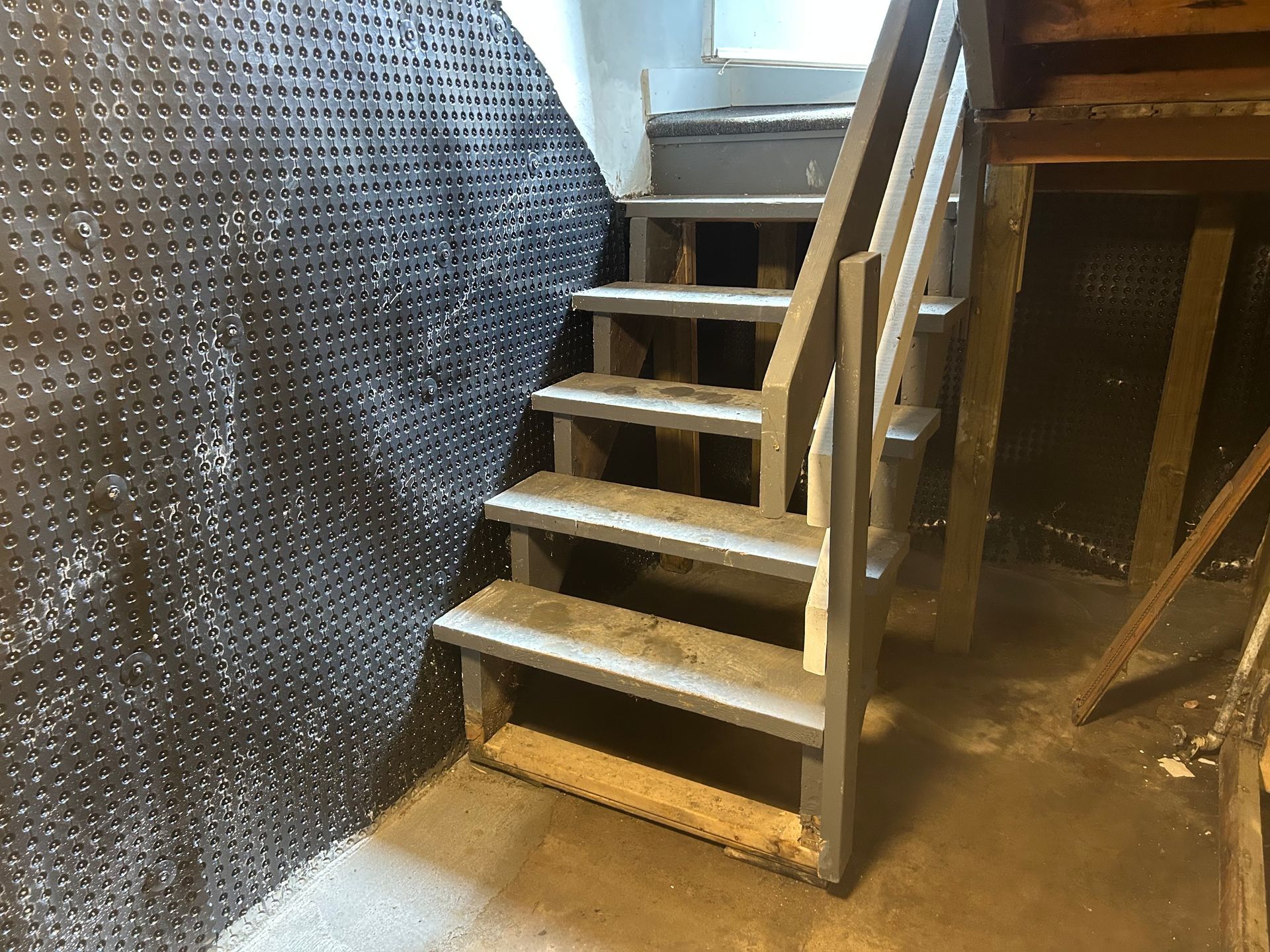 Wooden stairs in a basement, gray and white with a handrail. Black textured wall on the left.