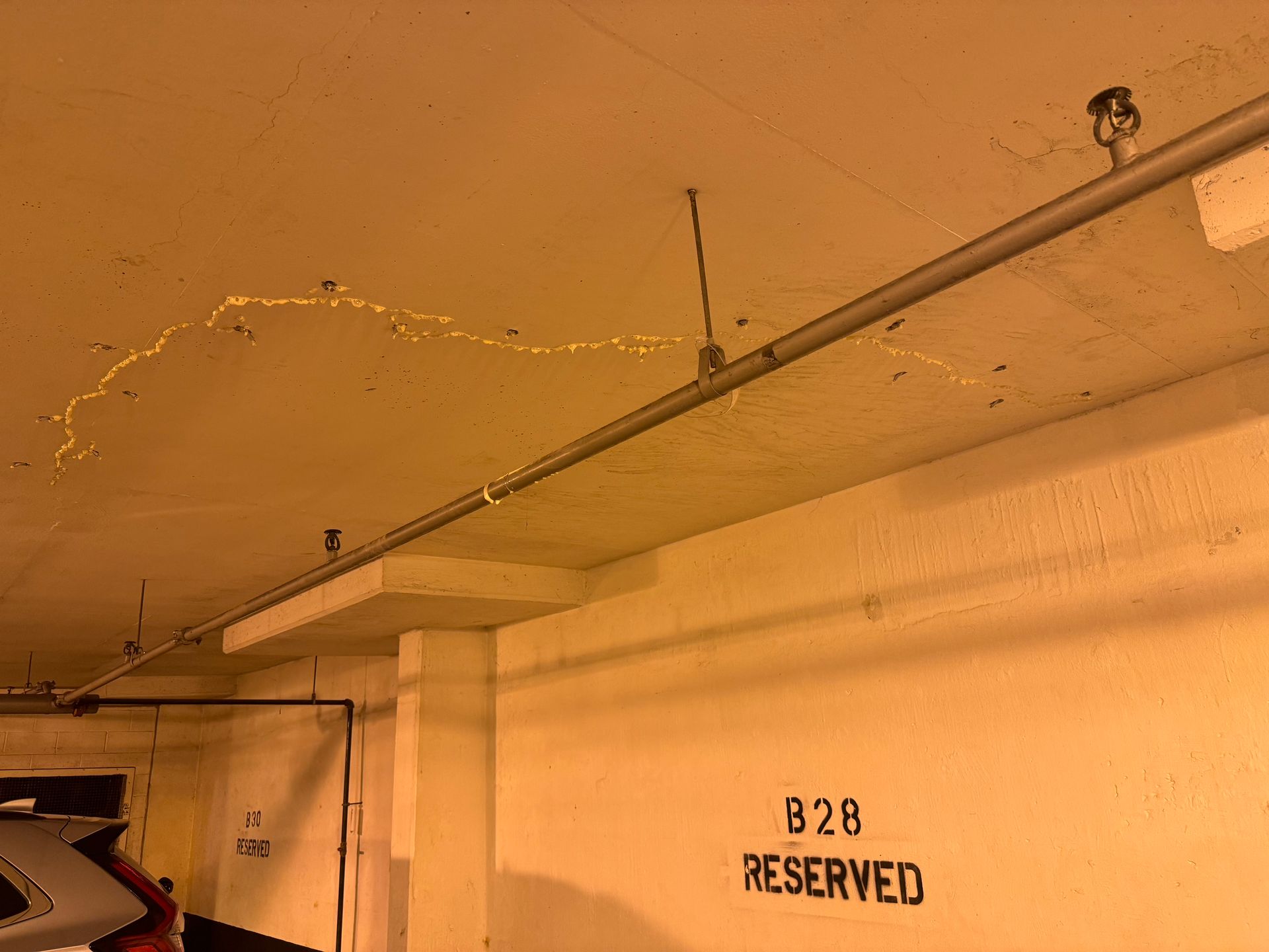 Yellow-tinted parking garage ceiling with water stains. A sprinkler system pipe is visible, as well as a 