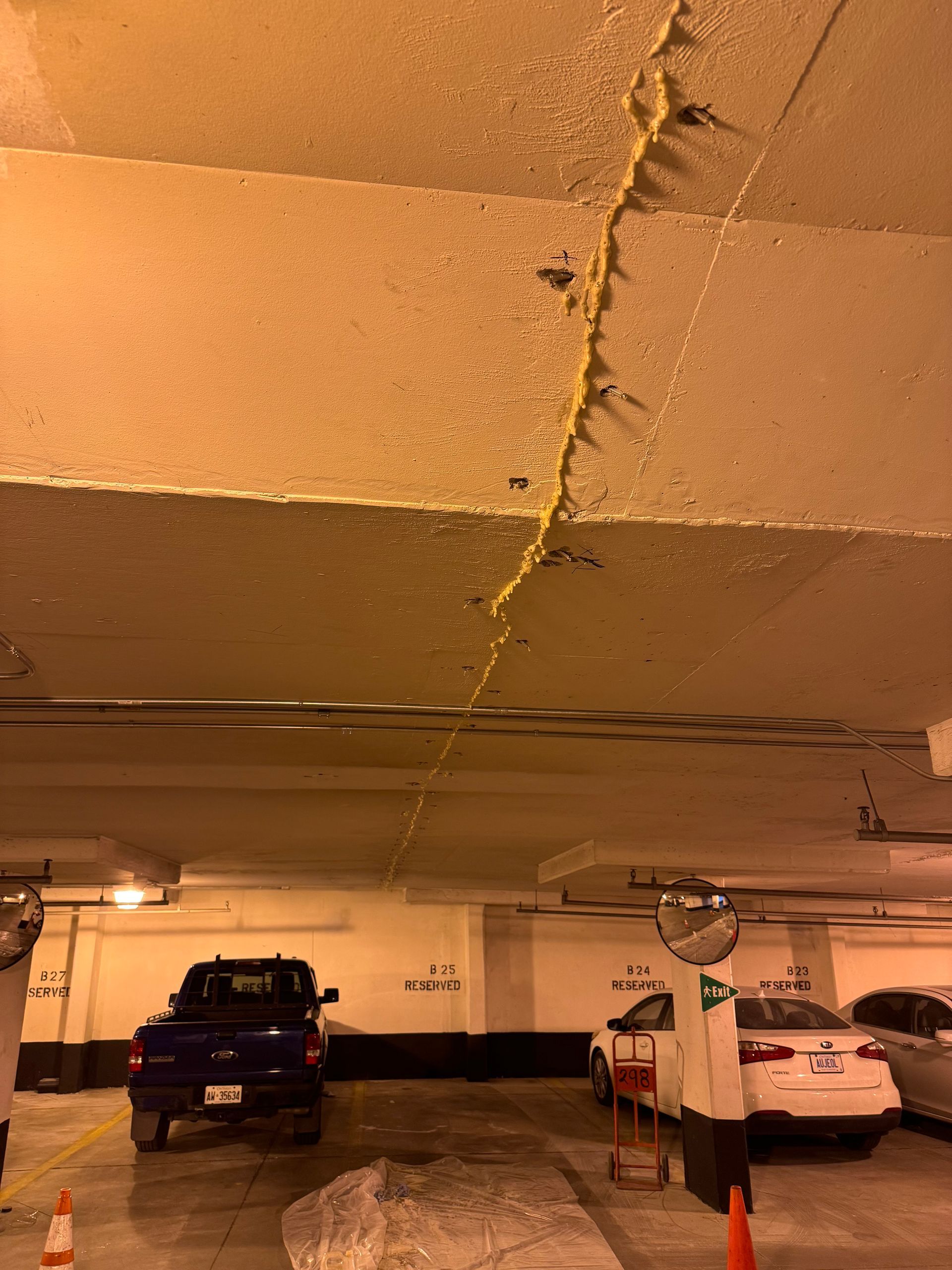 Crack in the concrete ceiling of an underground parking garage. Yellow sealant fills the crack. Cars parked below.