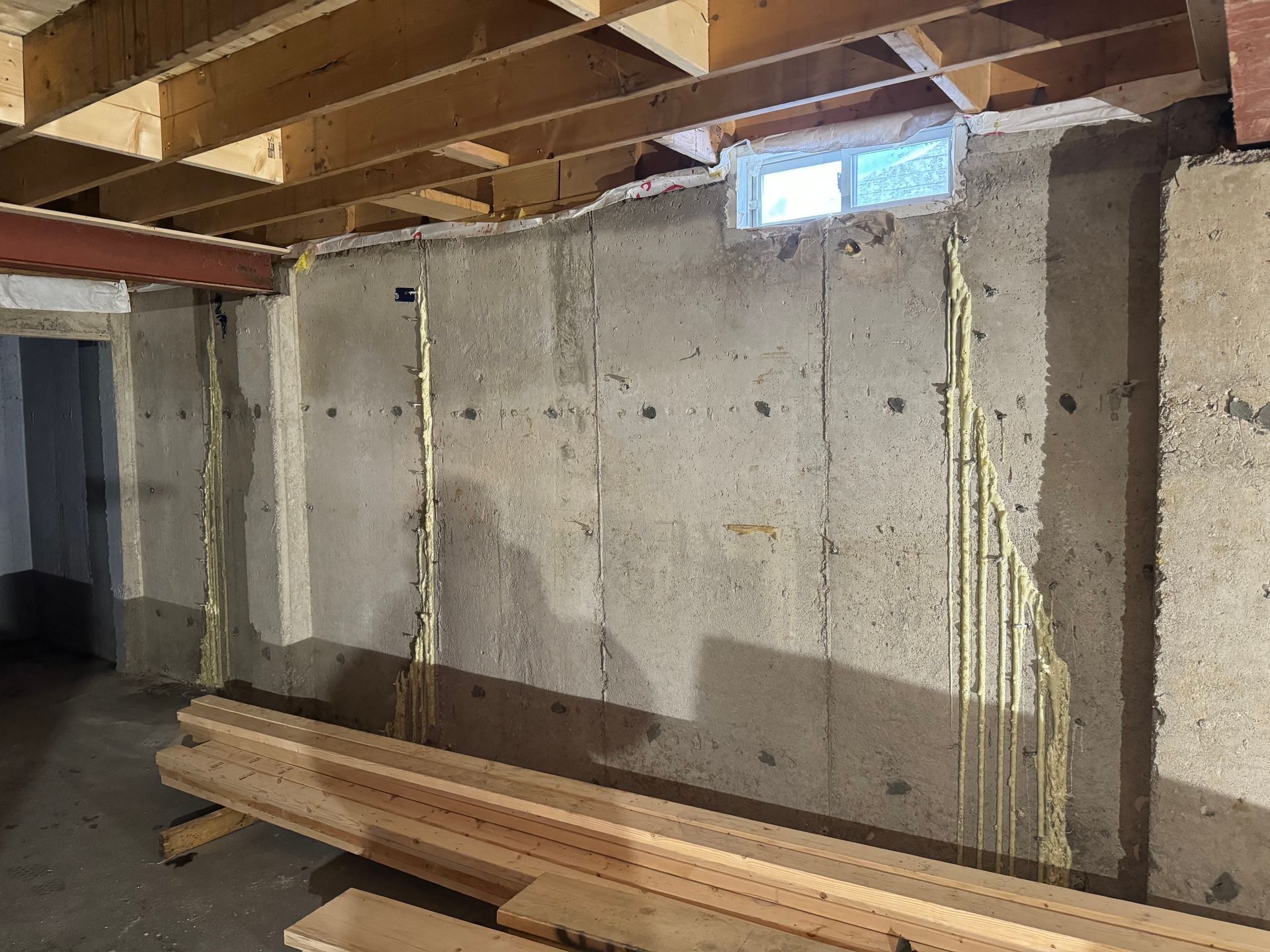 Basement wall with exposed electrical wiring and a small window. Wood beams are visible on the ceiling. Boards are stacked on the floor.