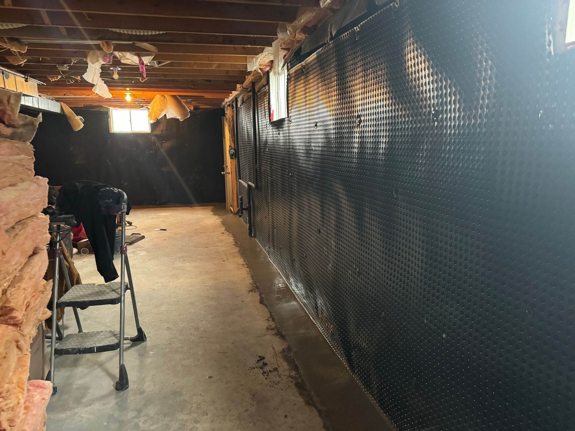 Basement interior with black dimpled wall membrane, concrete floor, step ladder, and exposed ceiling.