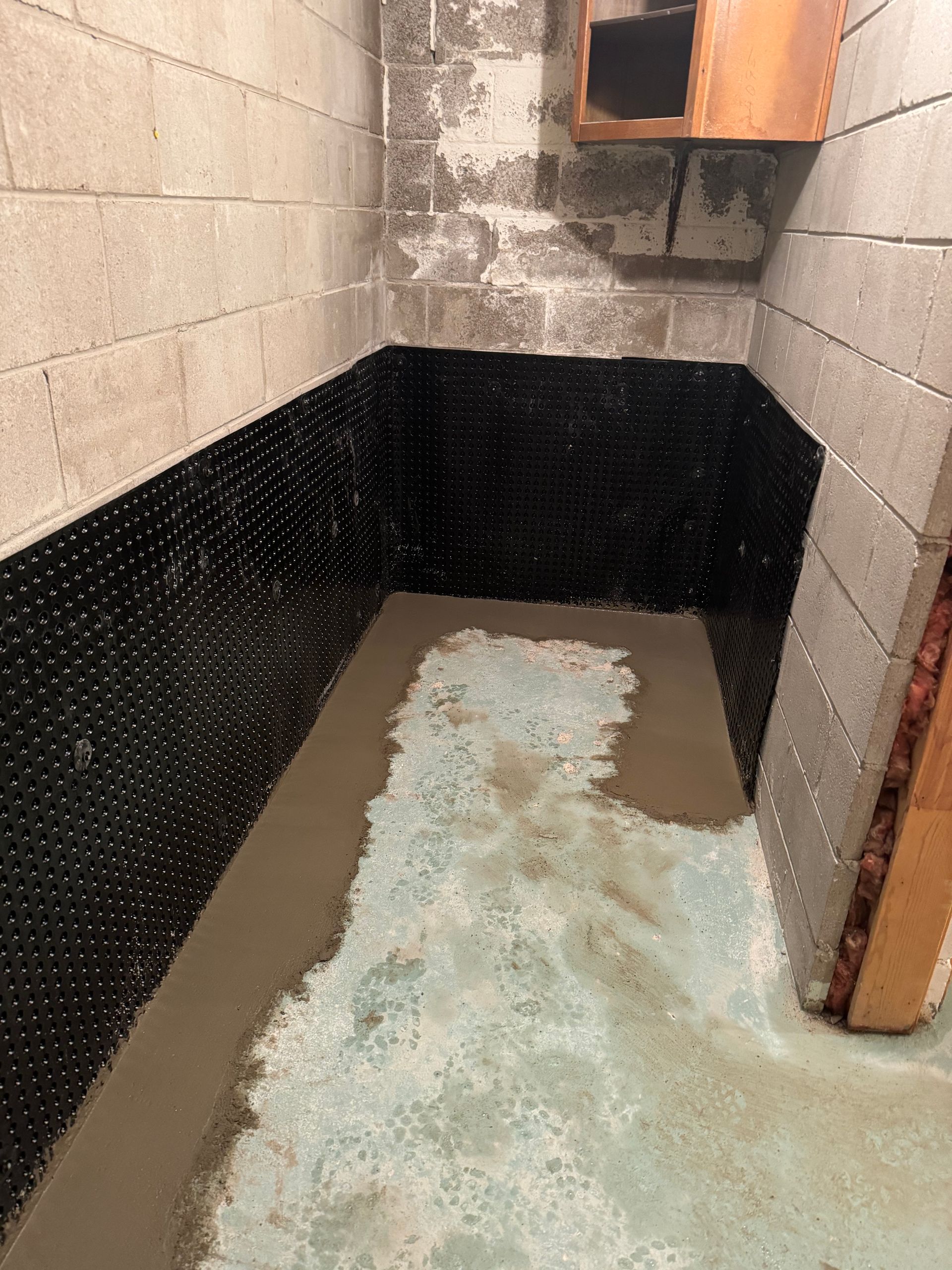 Basement corner with black waterproof membrane on walls, concrete floor with recent cement application.