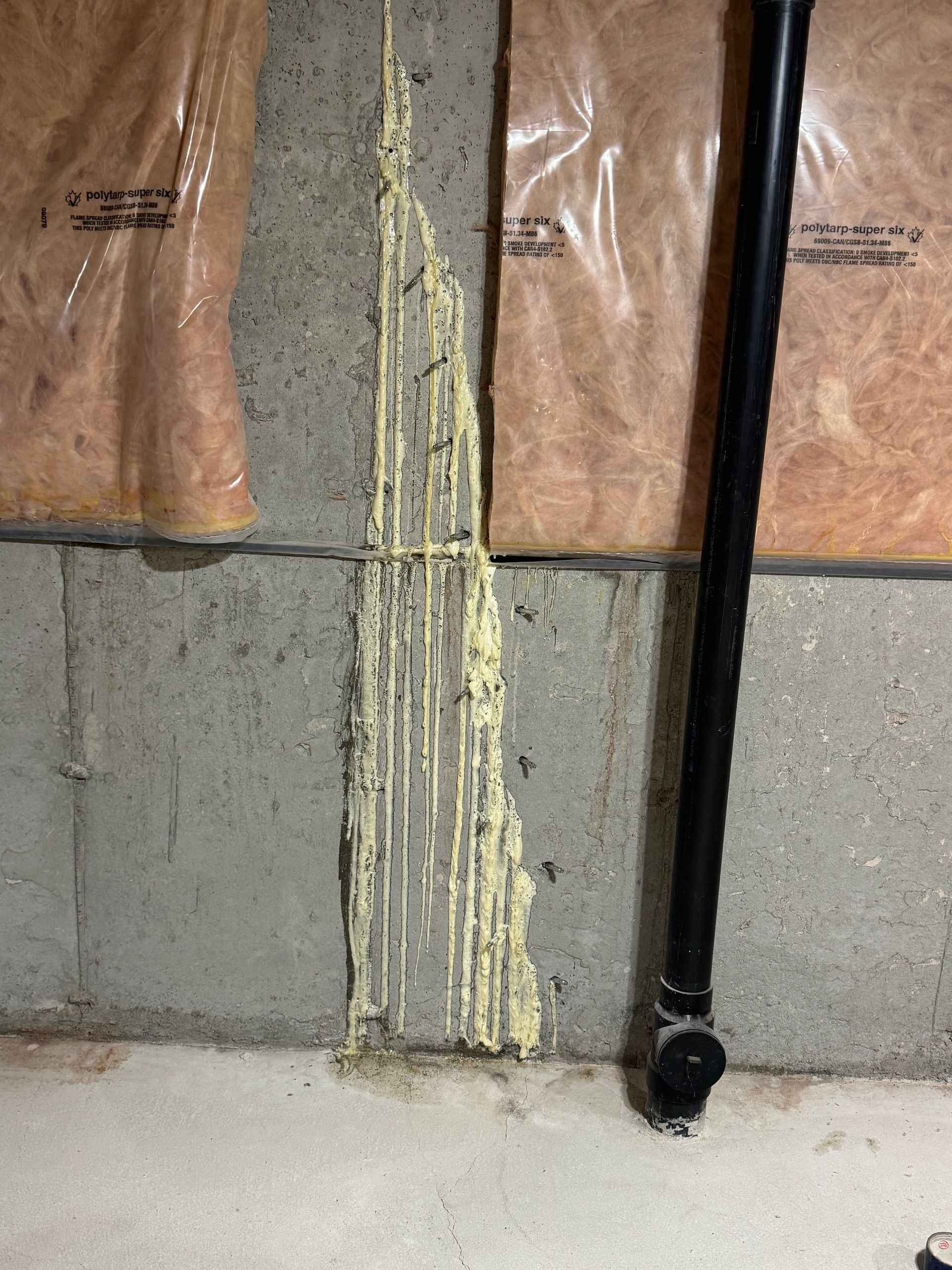 Gray concrete wall with yellow sealant. Black pipe and insulation present in a basement.