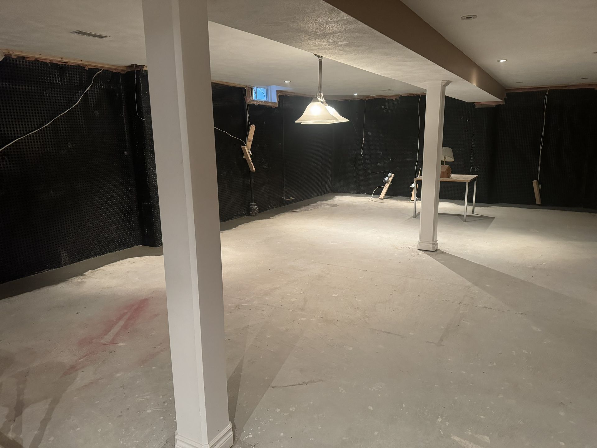 Empty basement with concrete floor, black insulation, and white support beams. A single hanging light fixture is visible.