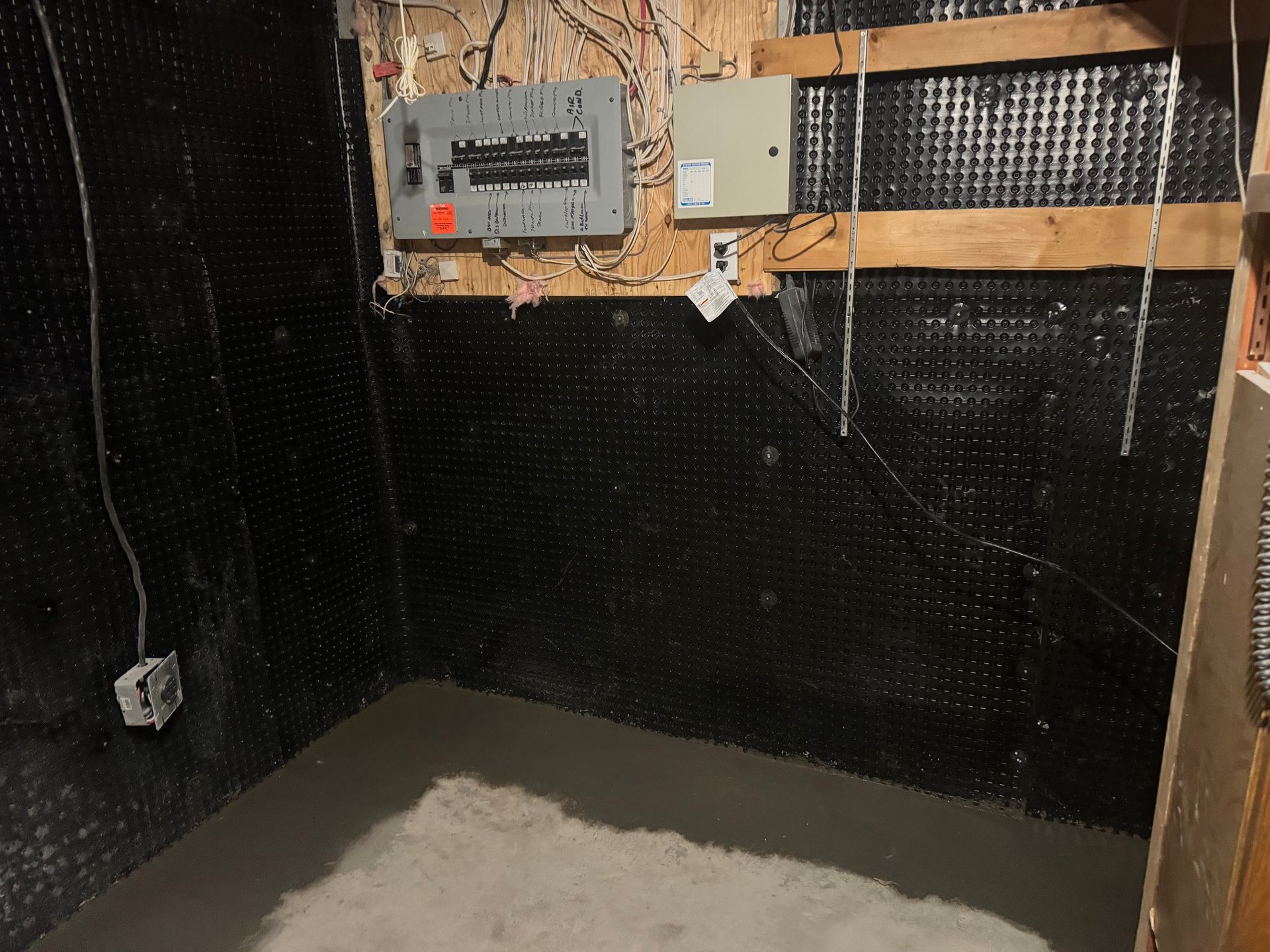 Basement interior with electrical panel, black waterproof membrane on walls, and a concrete floor.
