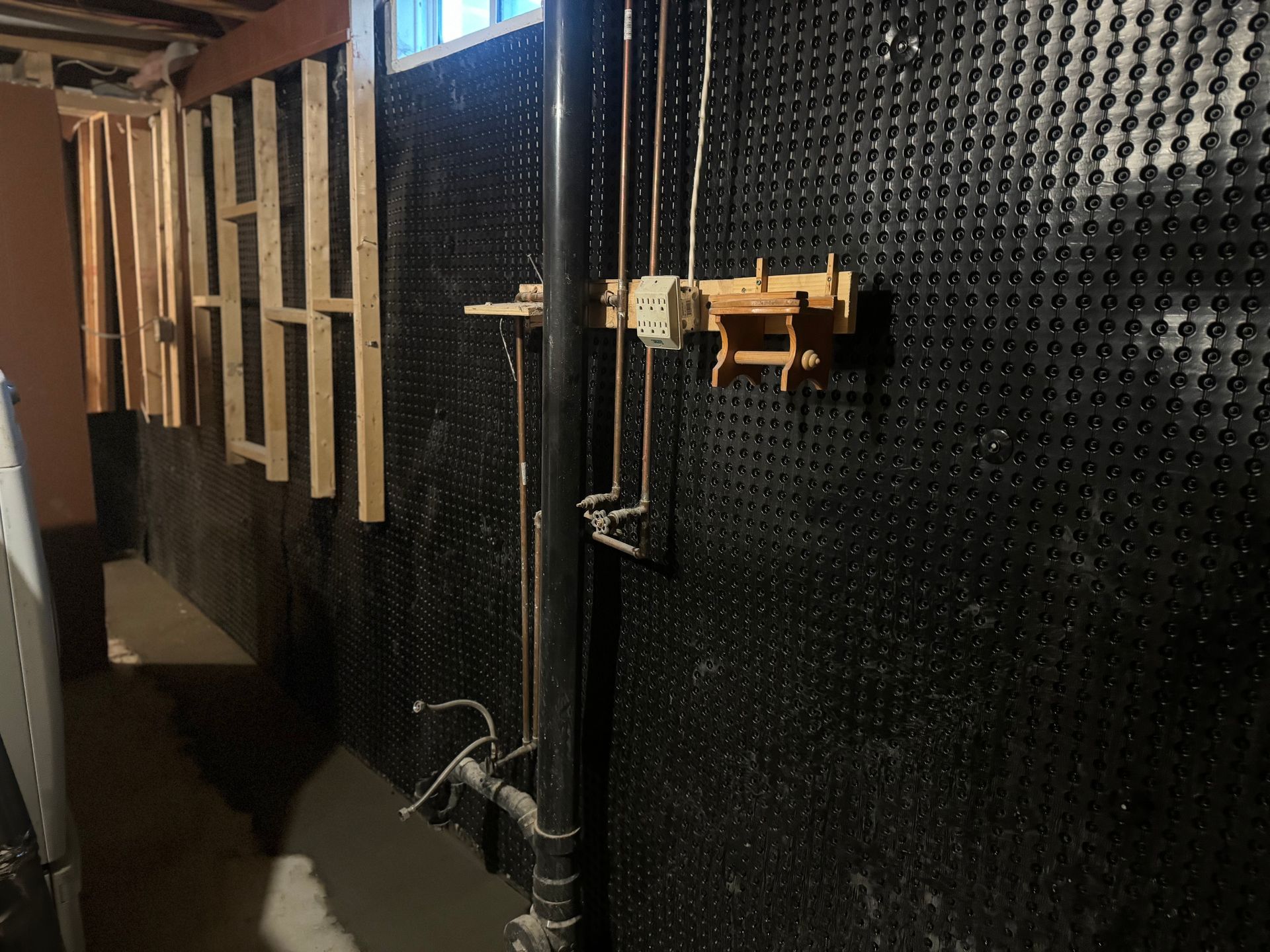 Black dimpled wall with pipes, framing, and an electrical box in a basement.