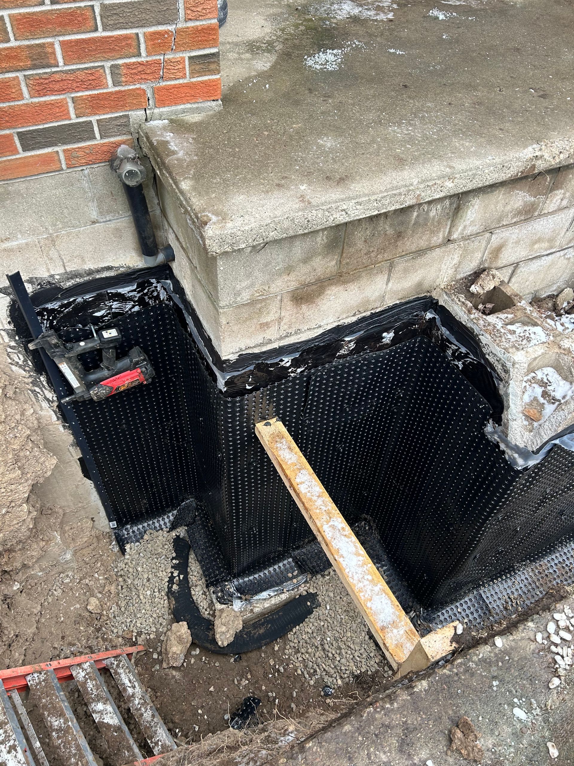 Black waterproofing membrane installed around concrete steps next to a brick wall.