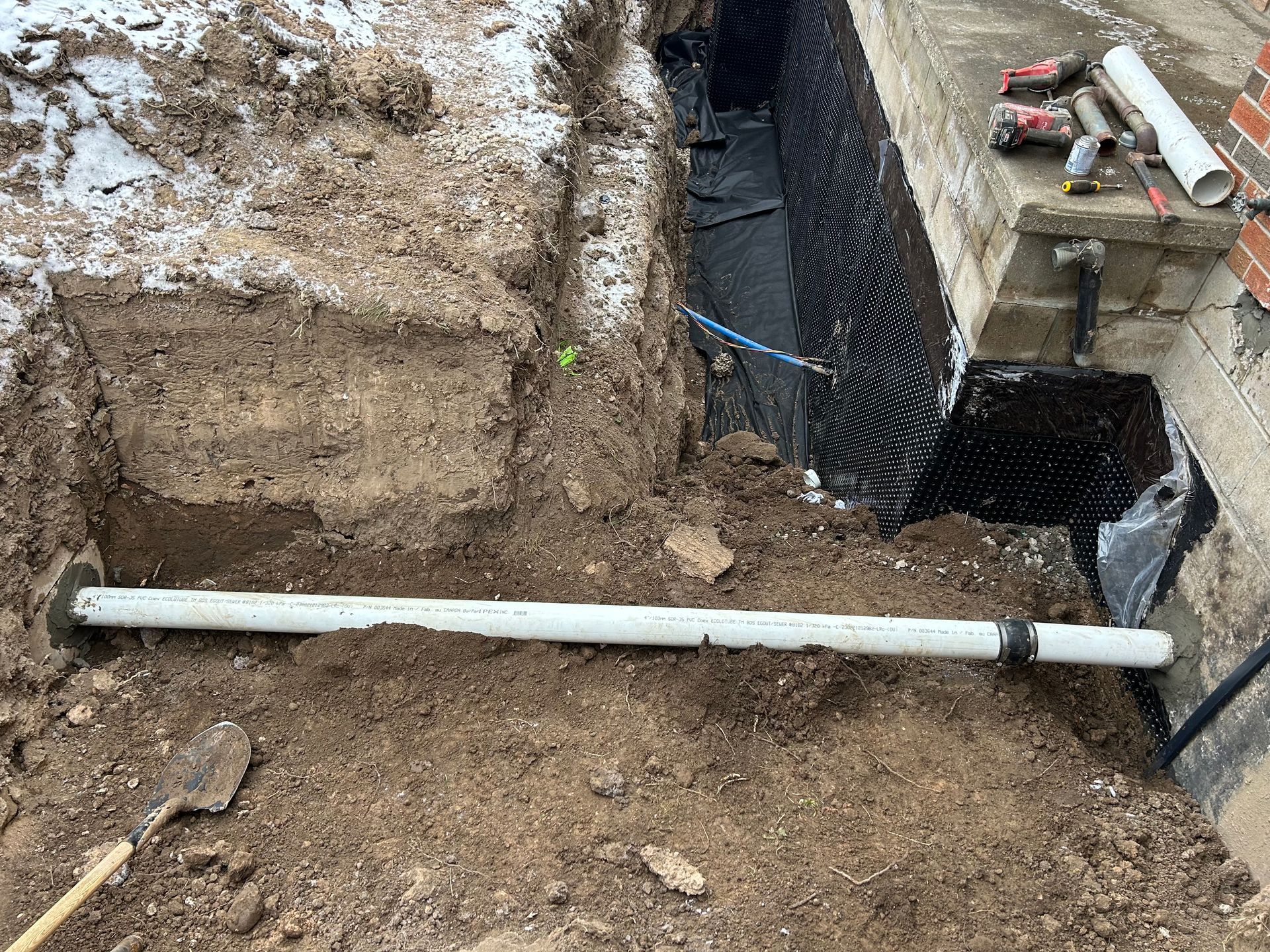 Trench dug along a brick wall, containing a white PVC pipe. Dirt and snow surround the pipe.