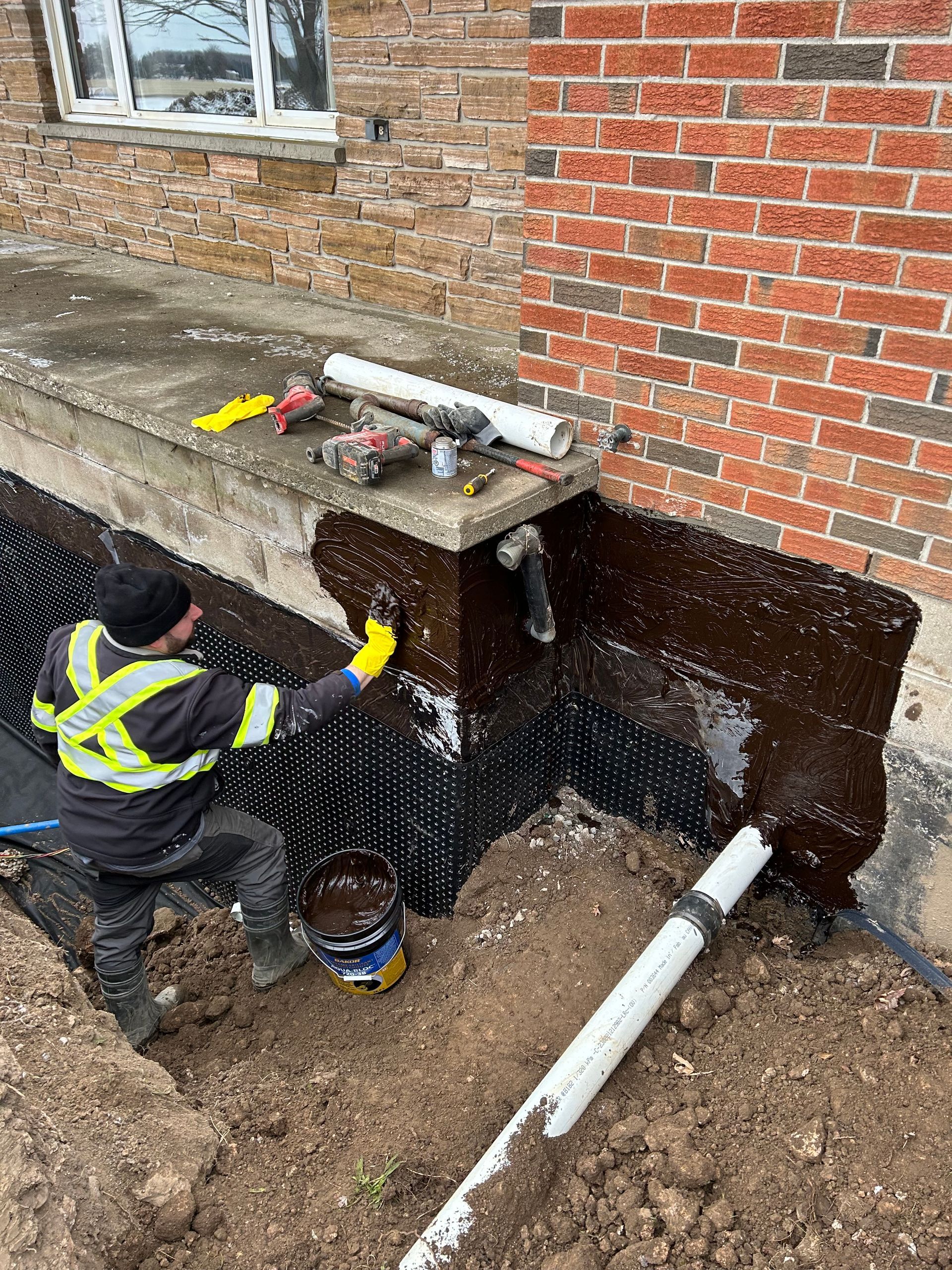 Man applying sealant to foundation with drainage membrane. Brick building and dirt.