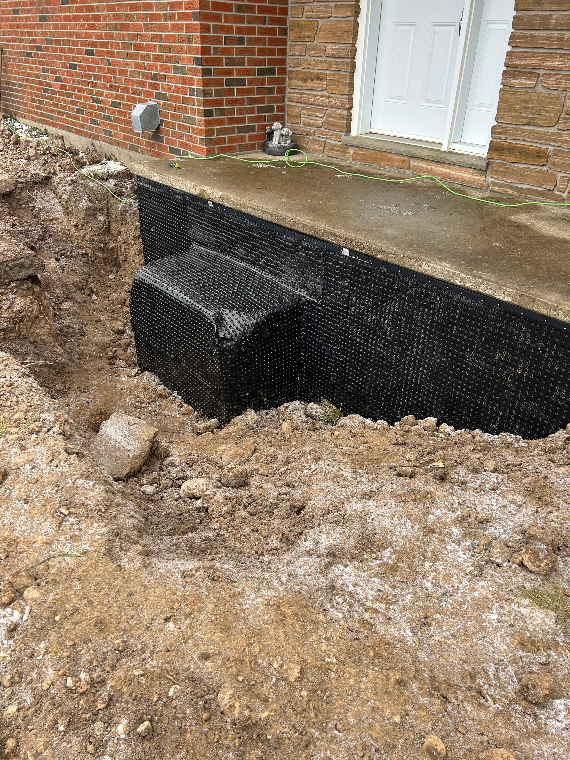 A foundation wall wrapped in black material is exposed in a trench dug beside a brick building.