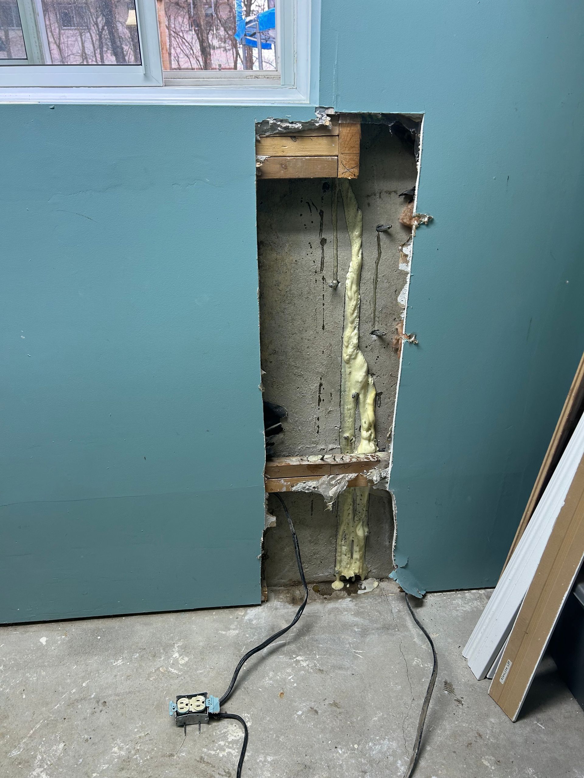 Hole in a teal wall, revealing concrete and wood framing, with exposed wires and foam sealant.