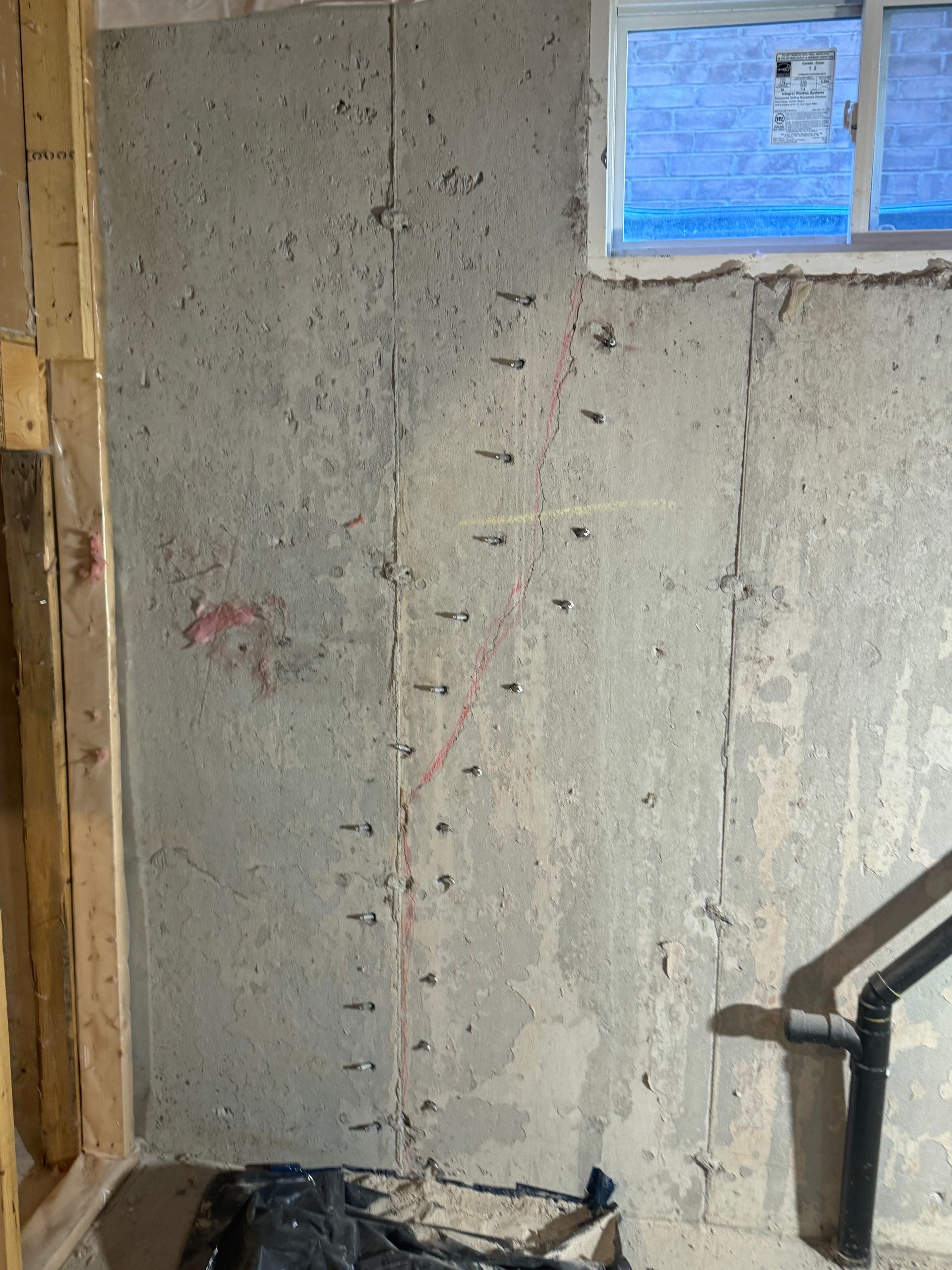 Concrete basement wall with exposed bolts, a window, and plumbing. Vertical seam with red markings.