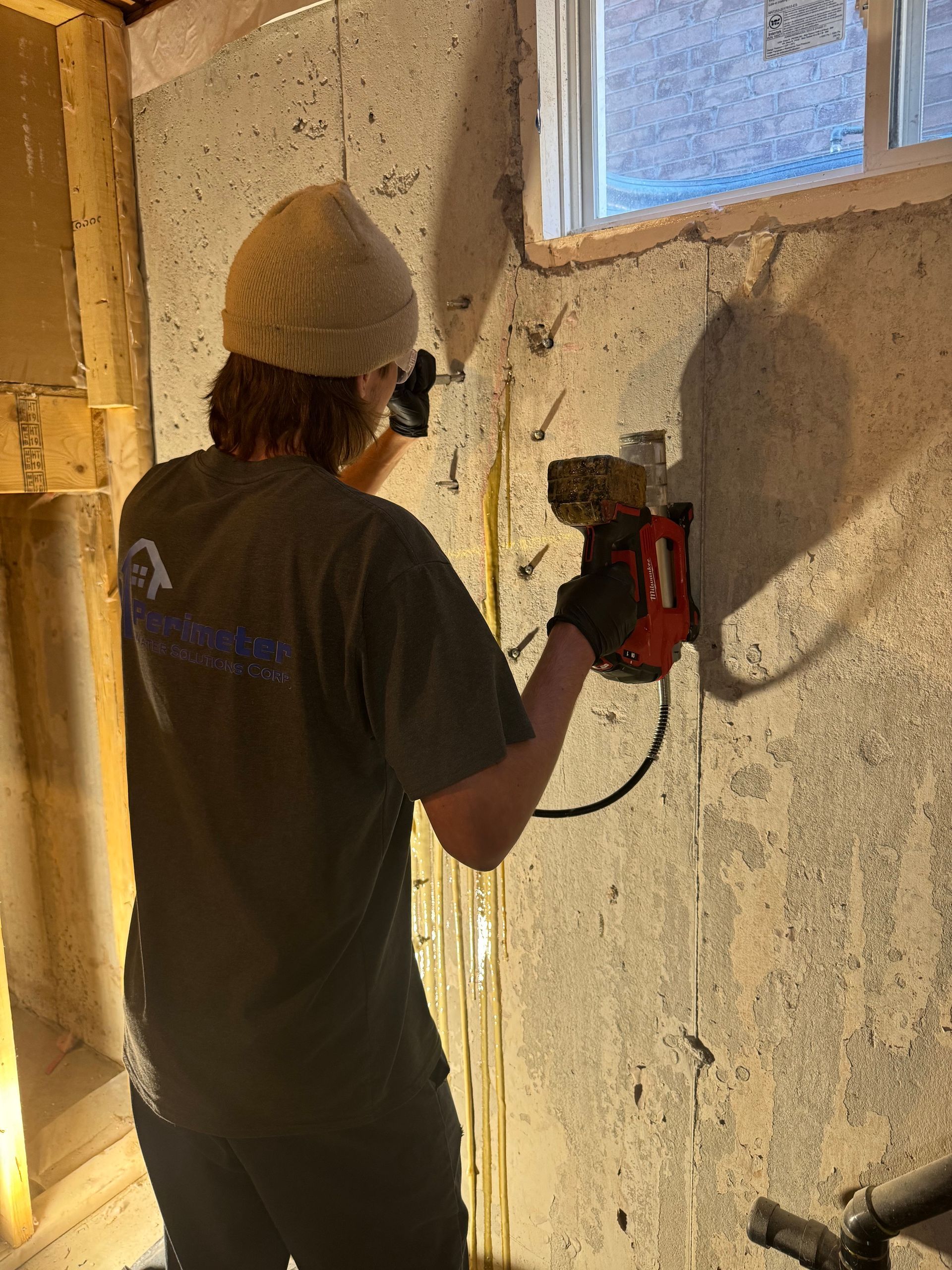 Person drilling a hole in a concrete wall, likely for electrical work. Wearing a beanie, holding a drill, in a basement.