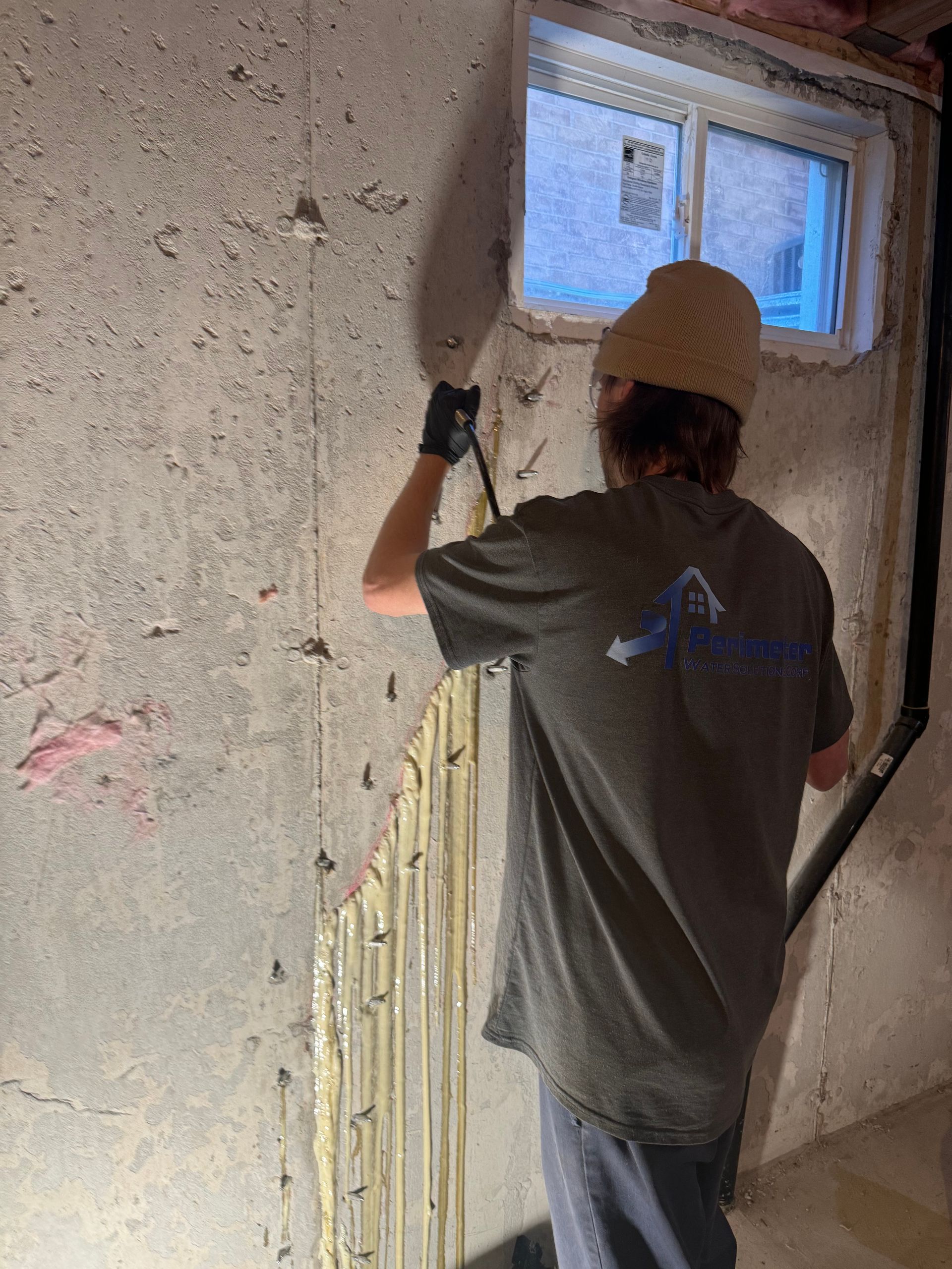 Person scraping a concrete basement wall near a window. They wear gloves, a hat, and a shirt with a logo.