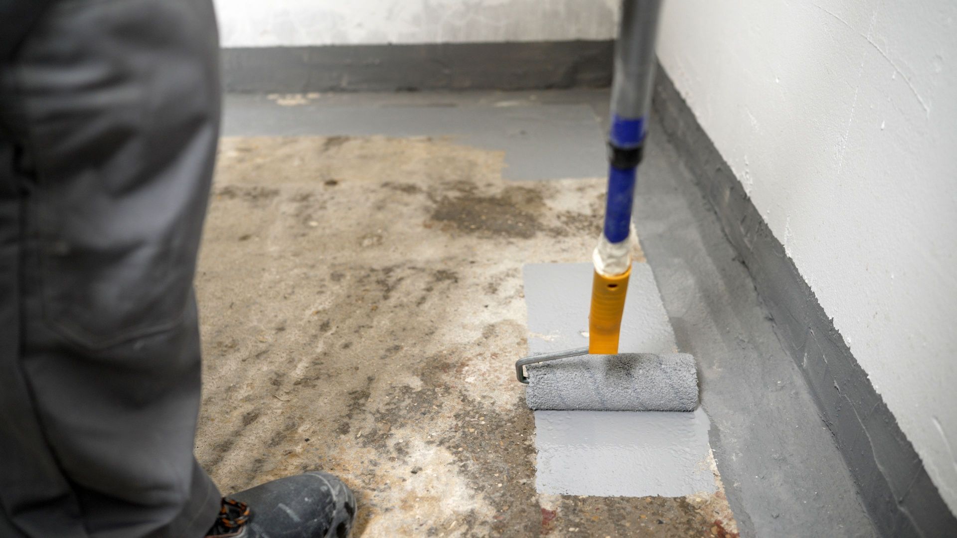 Person using a paint roller to apply gray paint to a concrete floor near a wall.