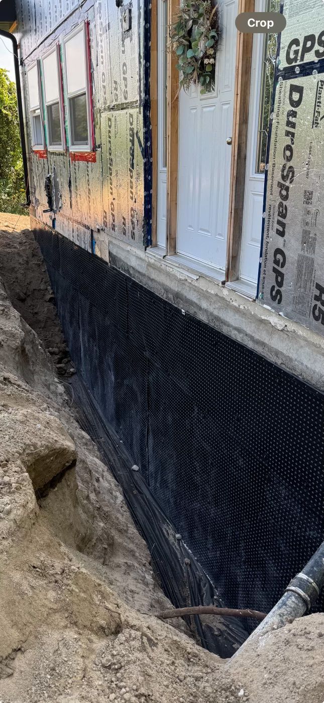 A building's foundation being worked on, with a trench dug around it. Black drainage material lines the foundation.