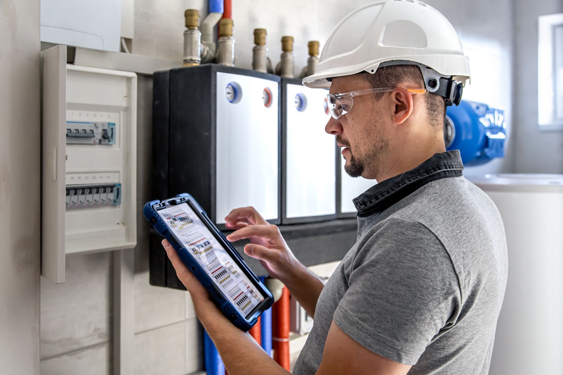 Electrician in hardhat, safety glasses, using tablet to inspect electrical panel.