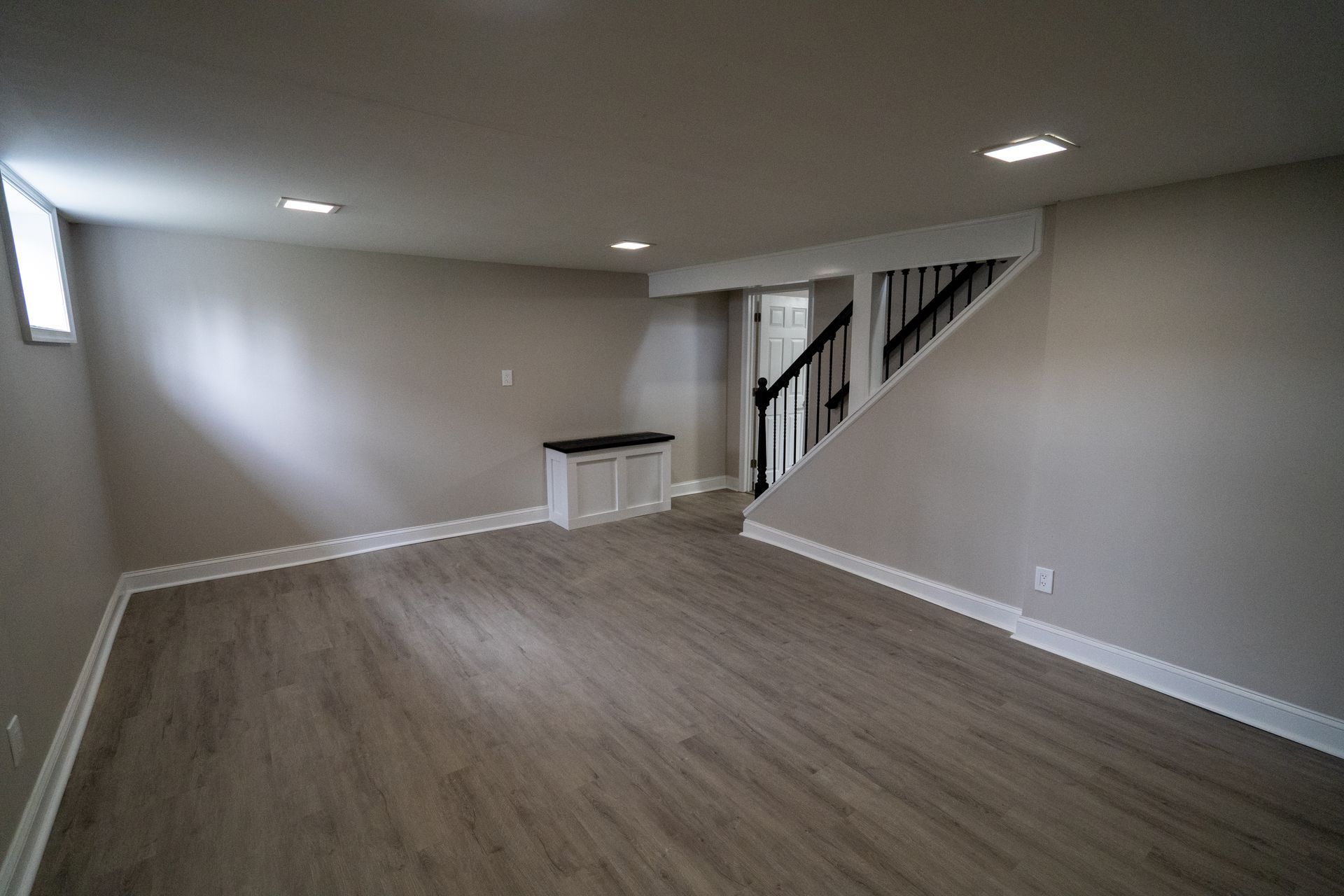 Empty basement room with light gray walls, wood-look flooring, and a staircase leading upwards.