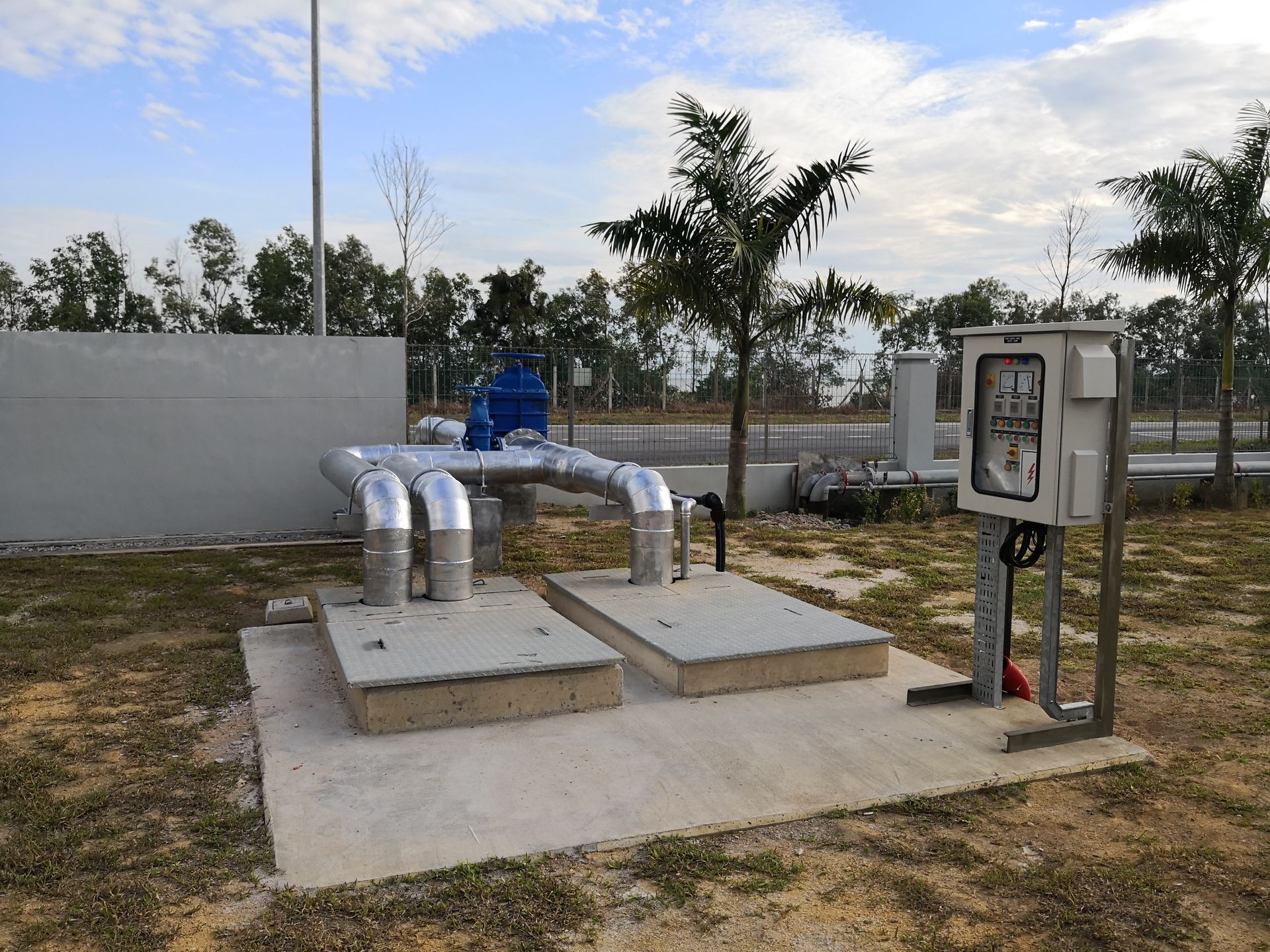 Outdoor utility infrastructure, pipes, and electrical box on a concrete pad with trees and a cloudy sky in the background.