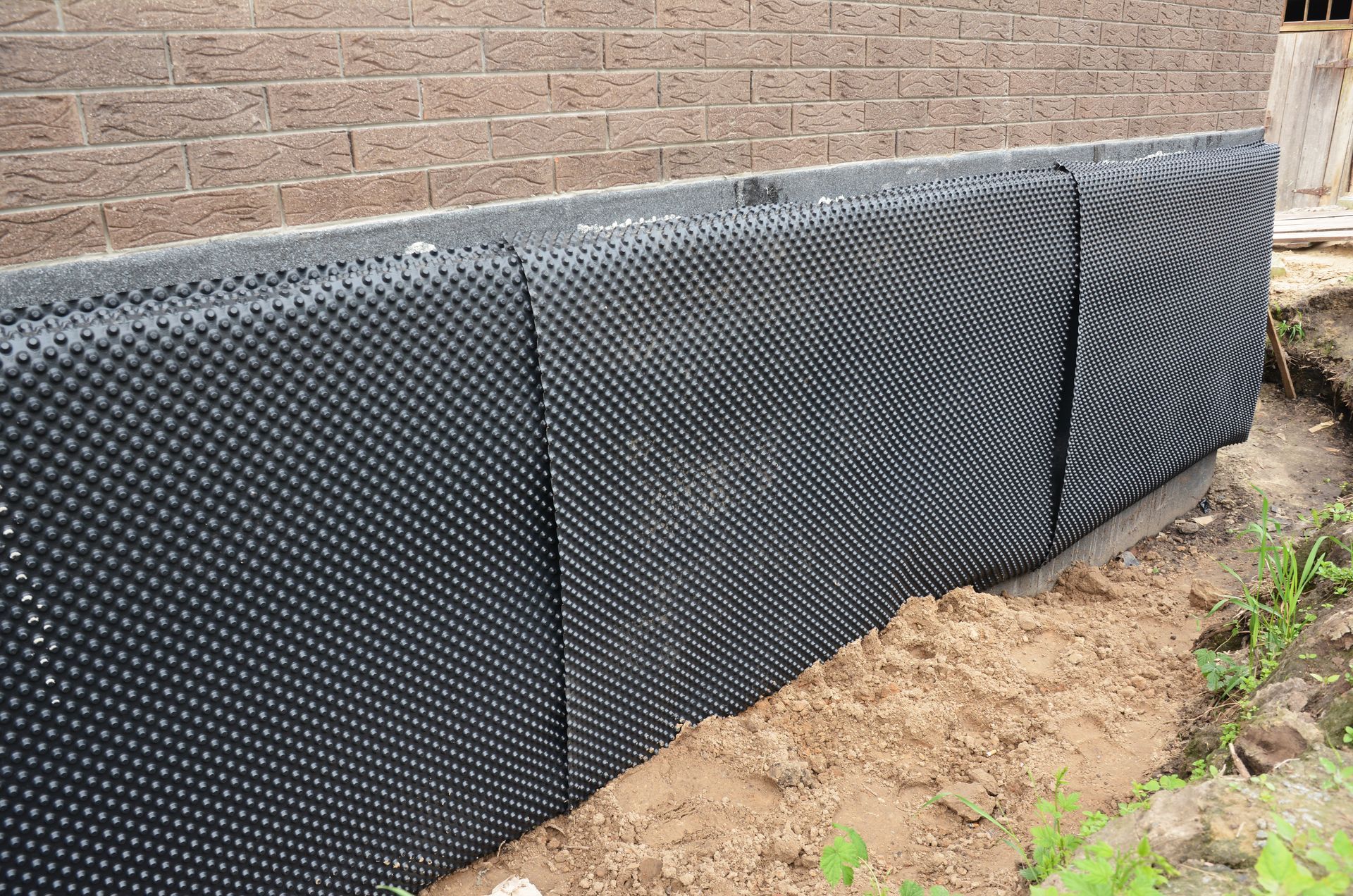 Black dimpled drainage membrane against a brick foundation, installed in an earthen trench.