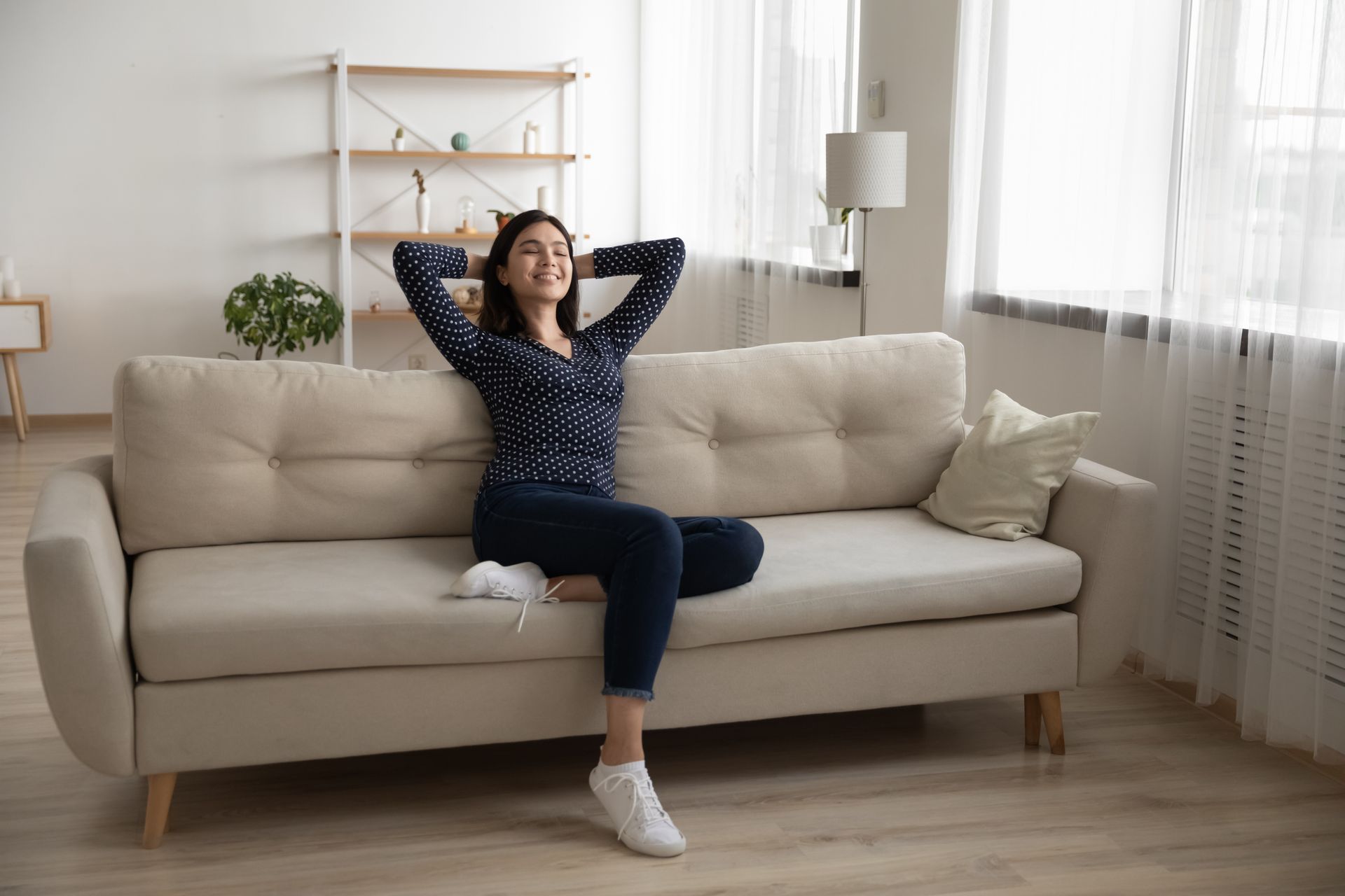 Woman relaxing on a beige couch, hands behind her head, smiling in a bright living room.
