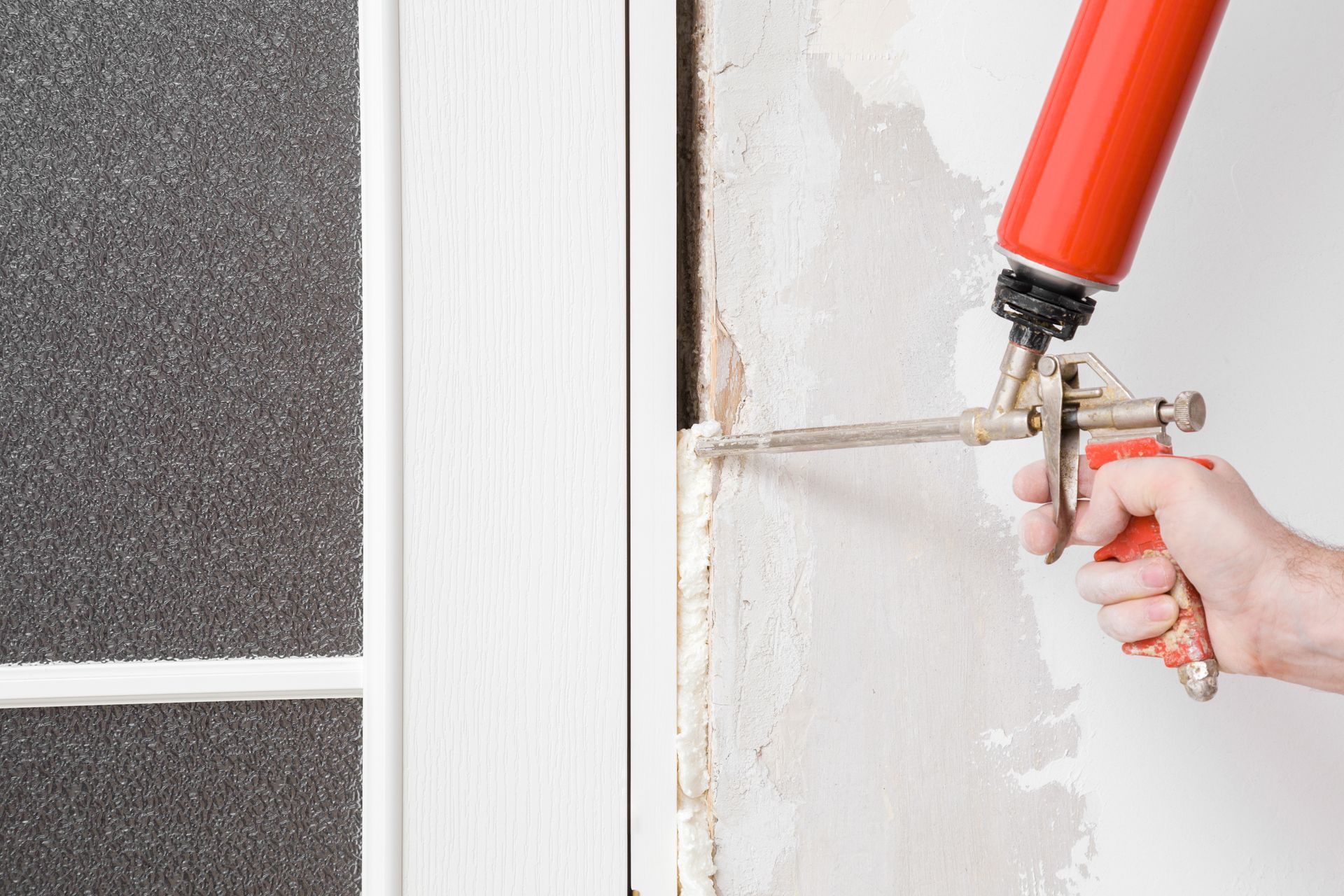 Hand using a red and gold foam gun to fill gaps around a white door frame.