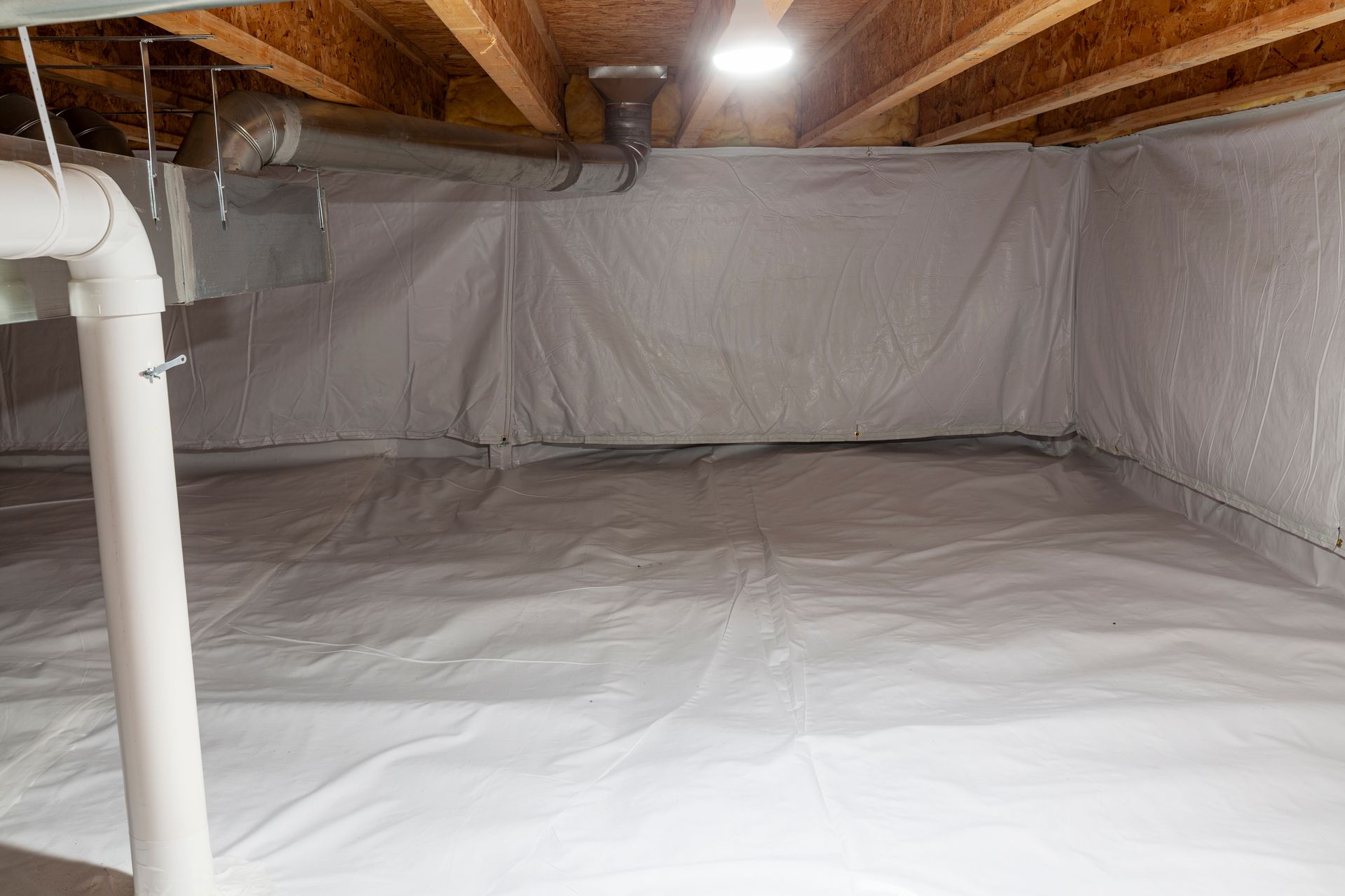 Crawl space encapsulated with white vapor barrier; exposed joists, PVC pipes, and light.