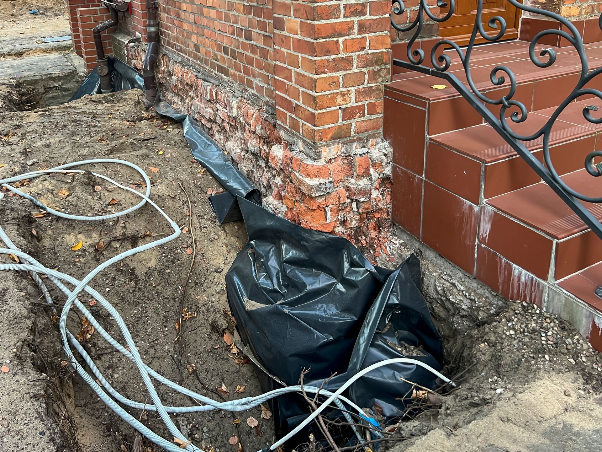 Excavated area next to a brick building. Black tarp, white tubes, and steps visible.