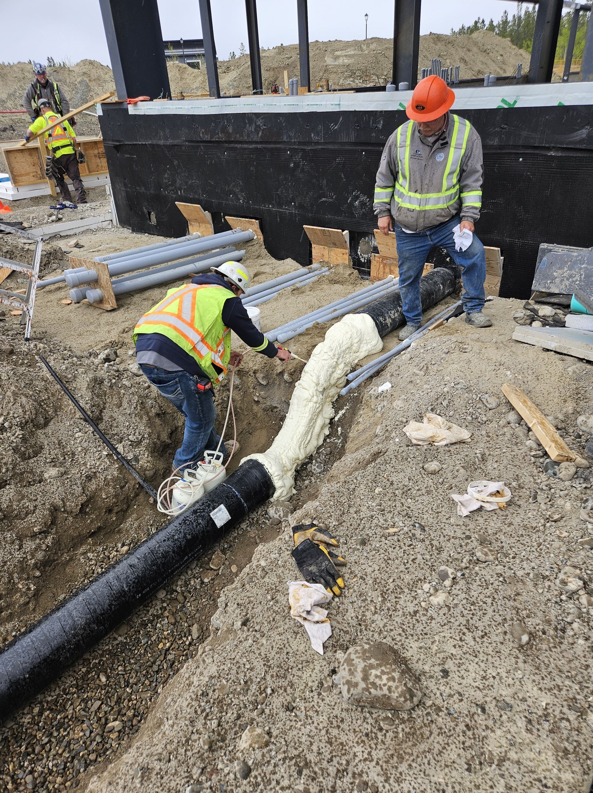 Construction workers applying insulation foam to a pipe in a trench at a construction site.