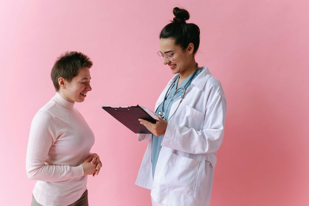 Doctor with clipboard smiles, talking with a patient against a pink backdrop.