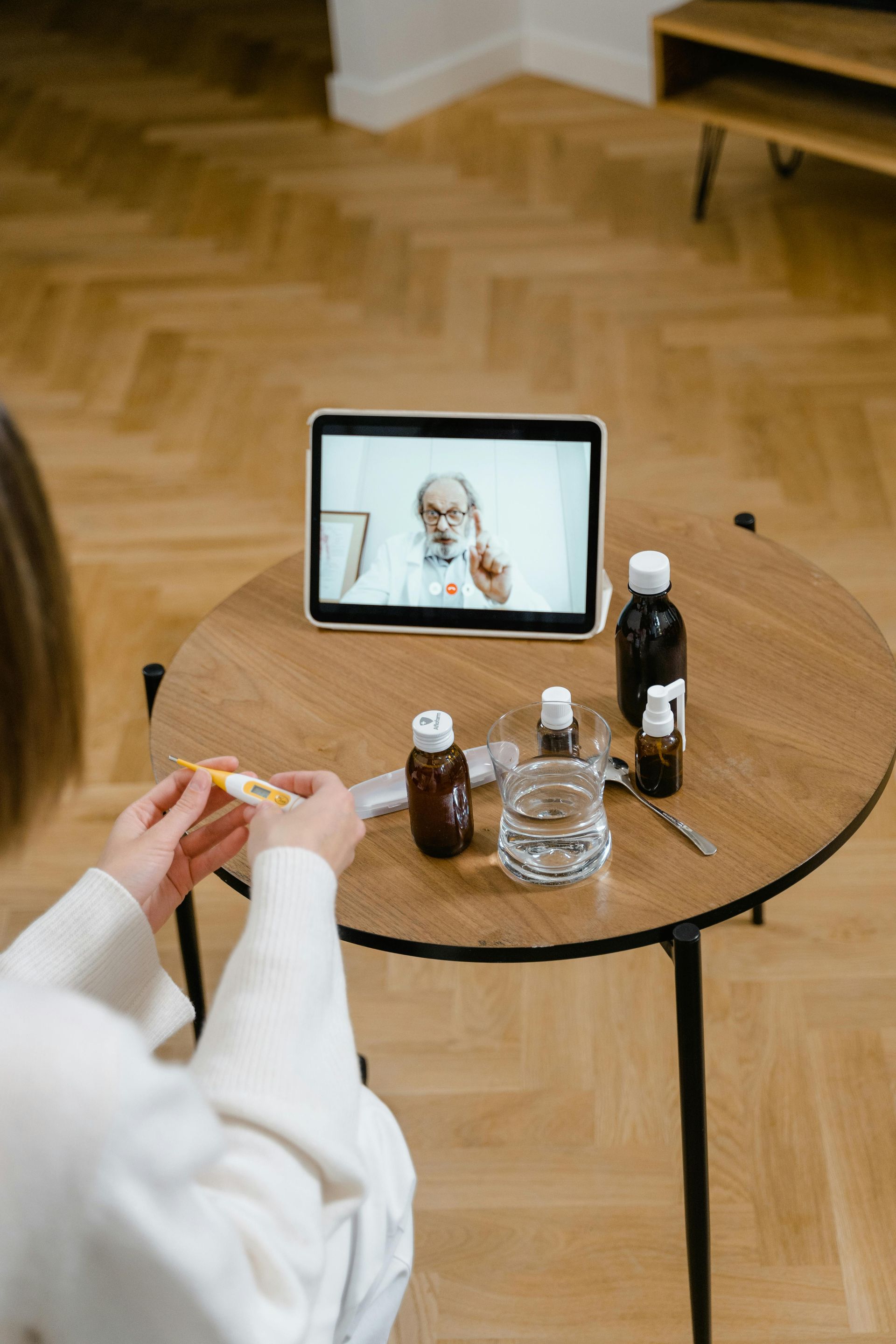 Woman in telemedicine appointment, holding thermometer, with doctor on screen and medicine on table.