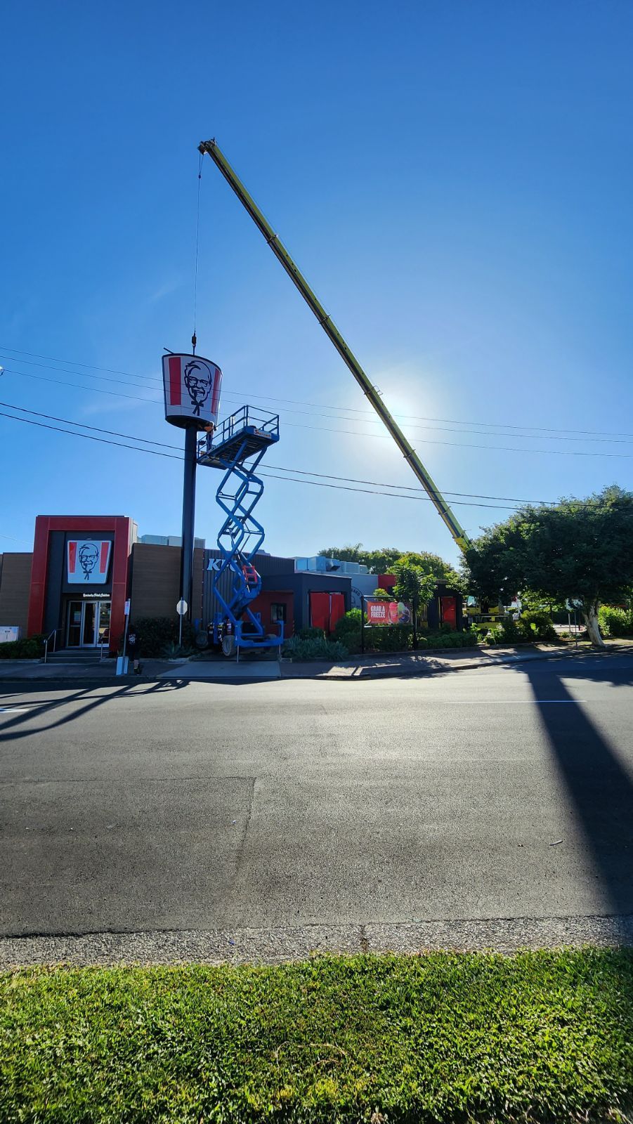 A KFC restaurant with a tall sign; a lift is by the sign. A long crane extends into the sunny blue sky — Statewide Lift & Shift Pty Ltd In Sarina, QLD