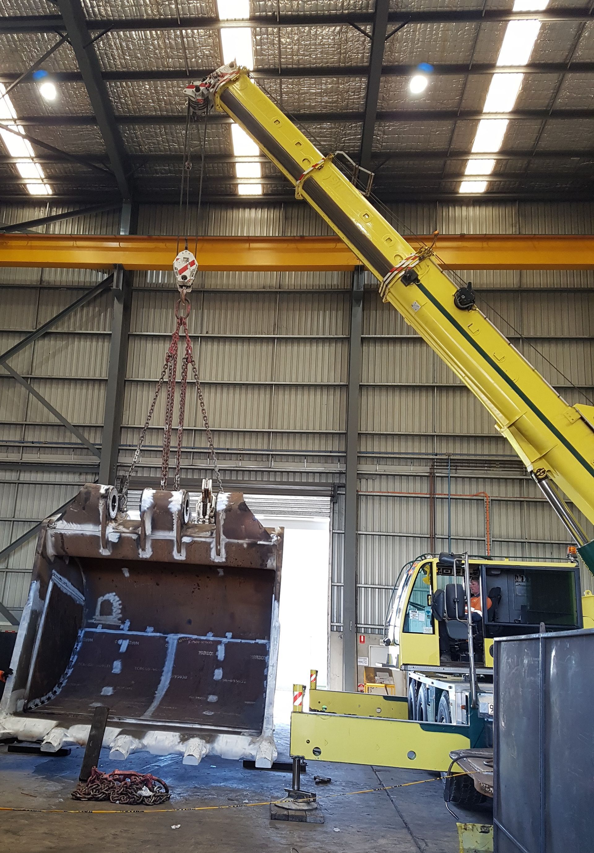 A yellow crane lifting a large, rusty excavator bucket inside a workshop — Statewide Lift & Shift Pty Ltd In Sarina, QLD