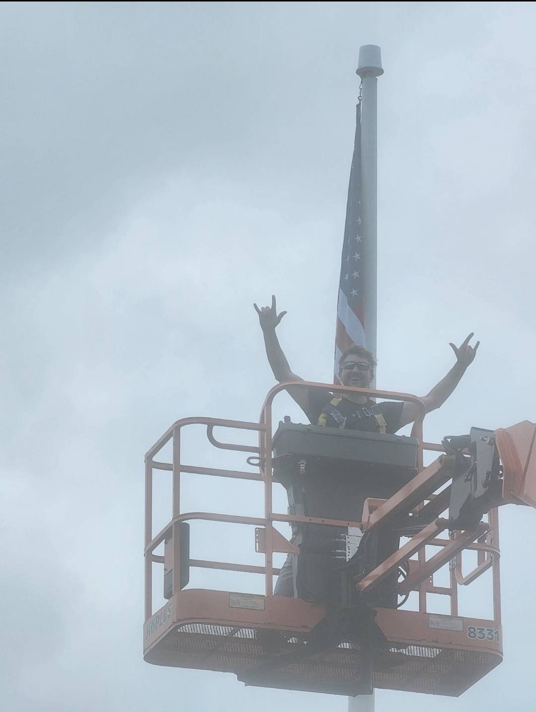 Man in lift basket raises arms while installing American flag on pole. Gray sky.