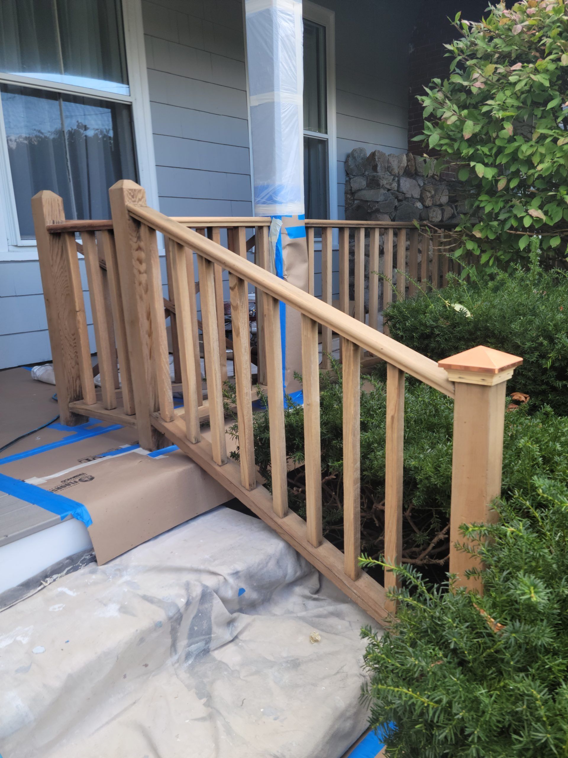 Wooden porch railing being prepped for painting, blue tape protecting adjacent surfaces.