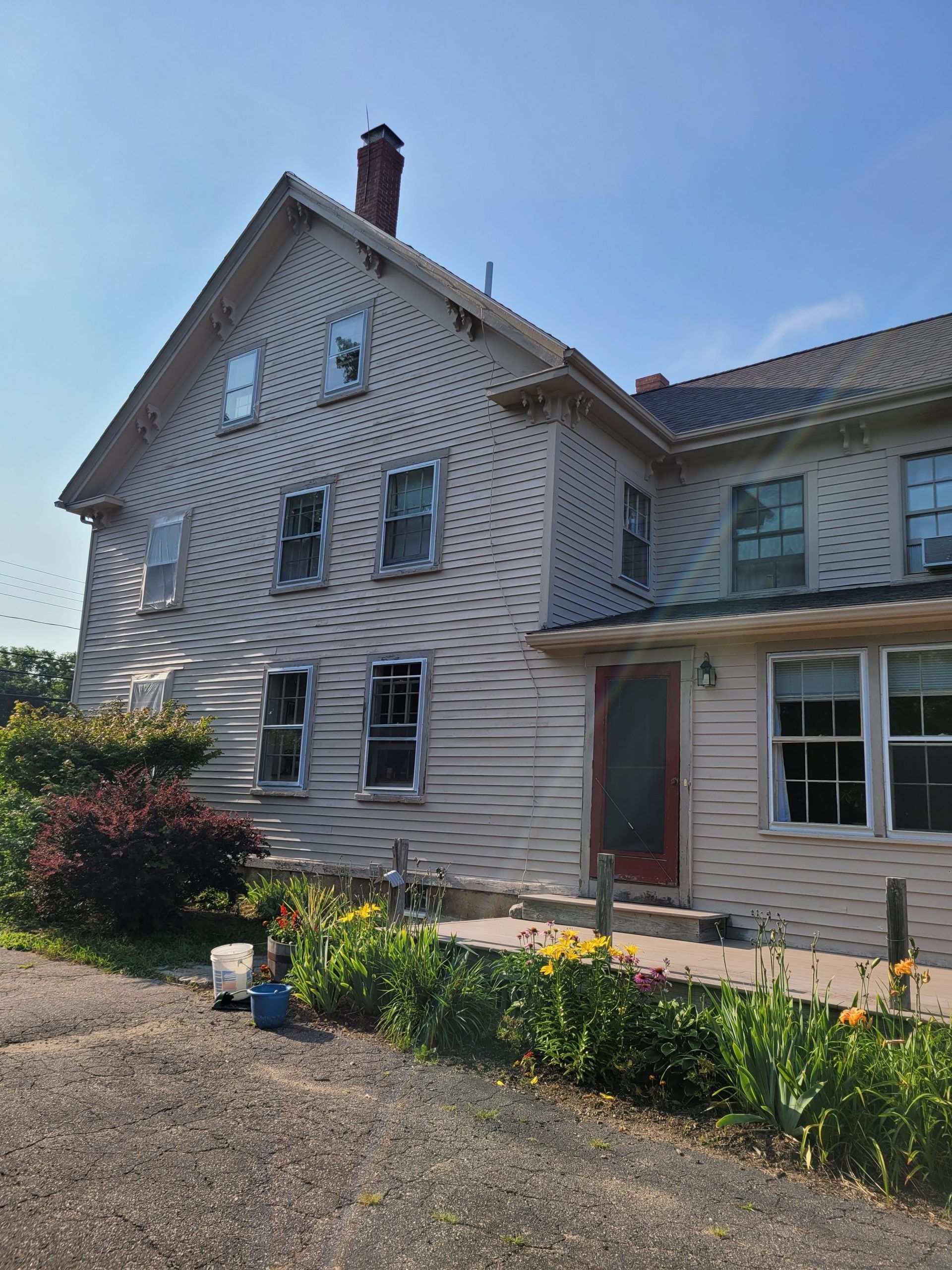 Two-story, light-colored house with multiple windows and a red door, surrounded by a garden, under a blue sky.