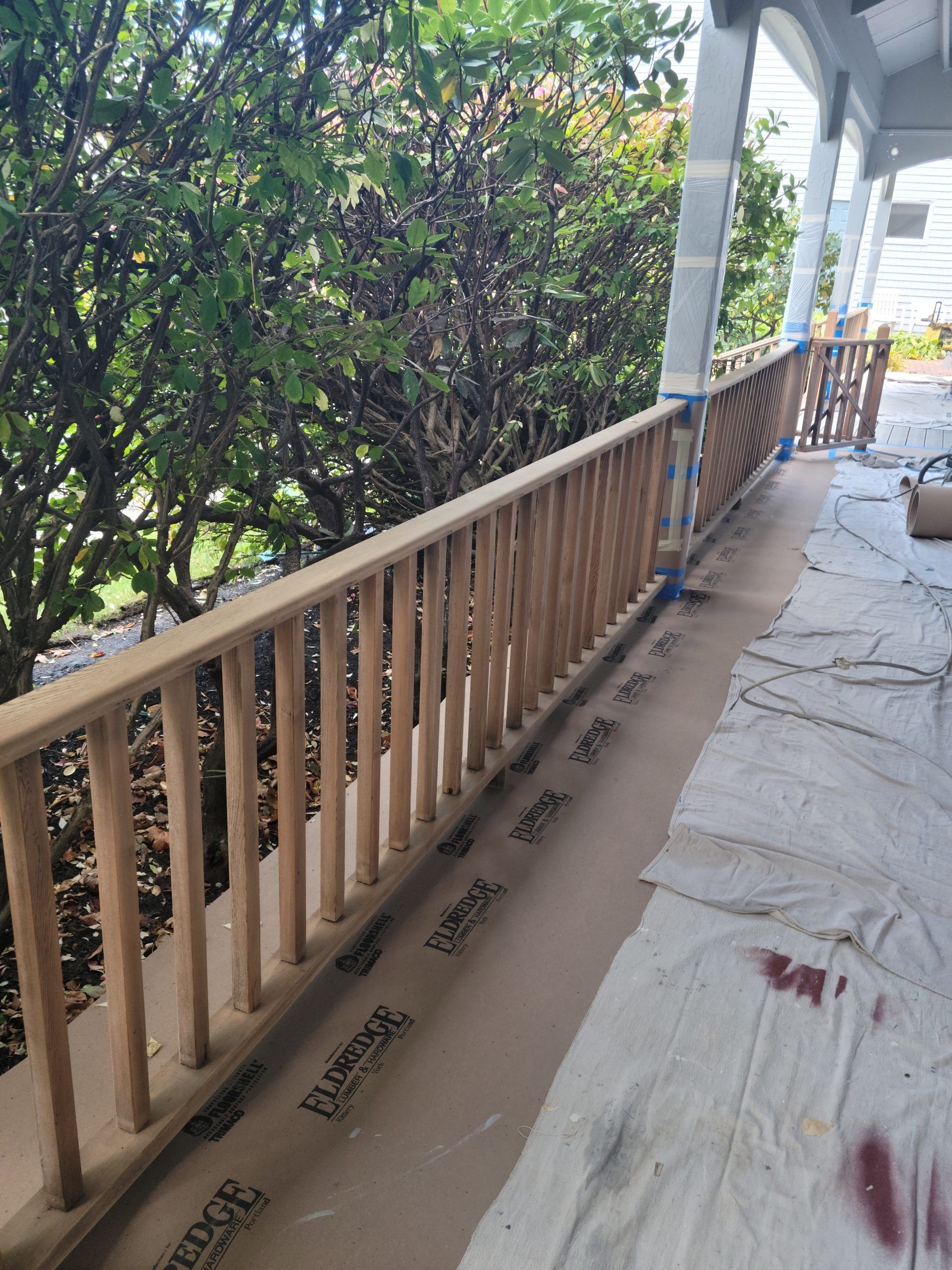 Wooden porch railing under construction, with blue tape, protective paper, and green foliage in background.