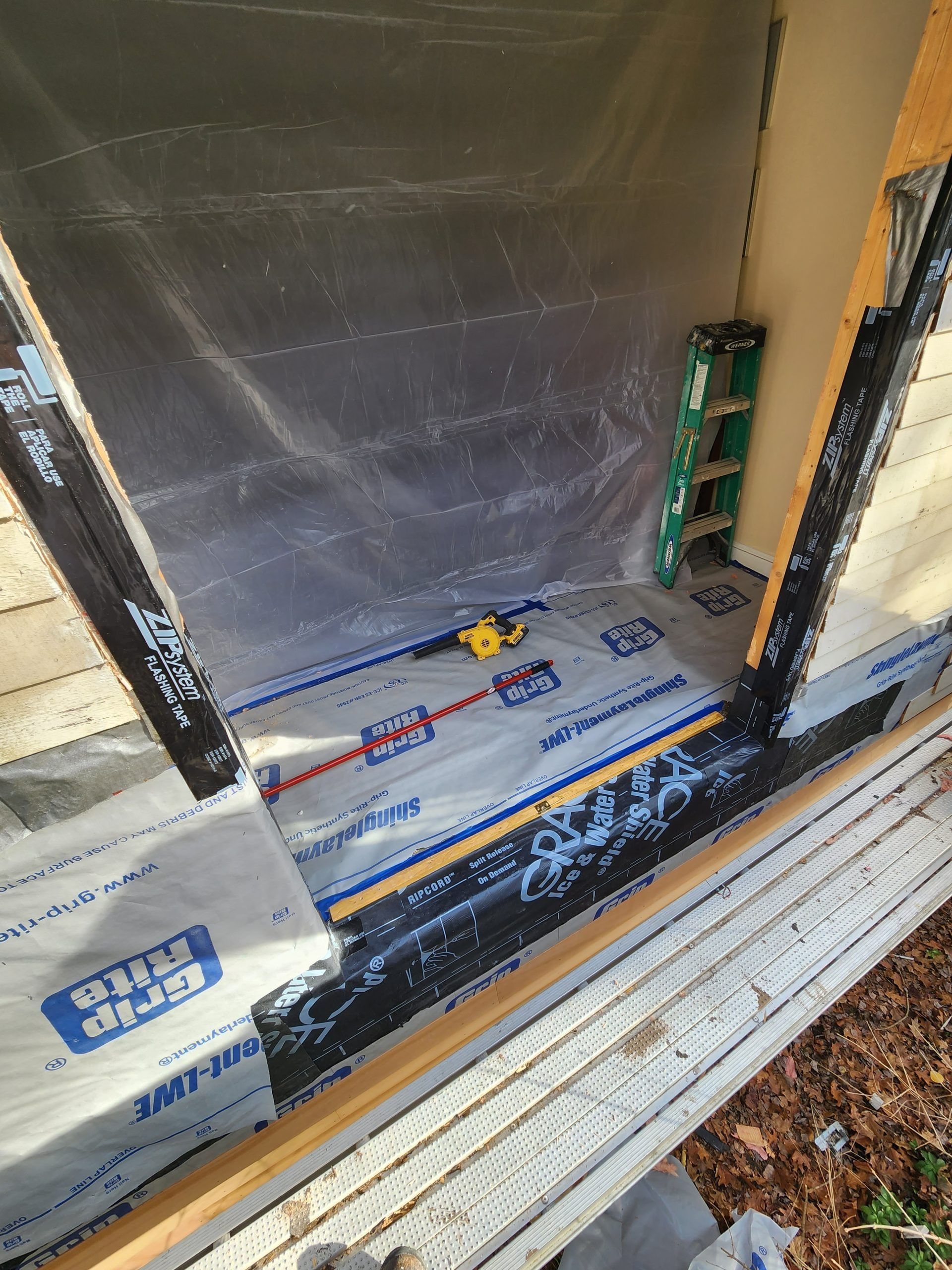Exterior doorway with black flashing tape, blue and white wrap, yellow tool, and a small green ladder.