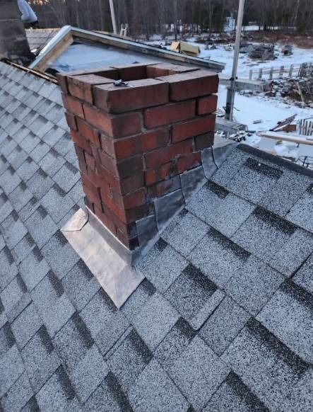 Brick chimney on a gray shingle roof, surrounded by flashing, with a partially snow-covered background.