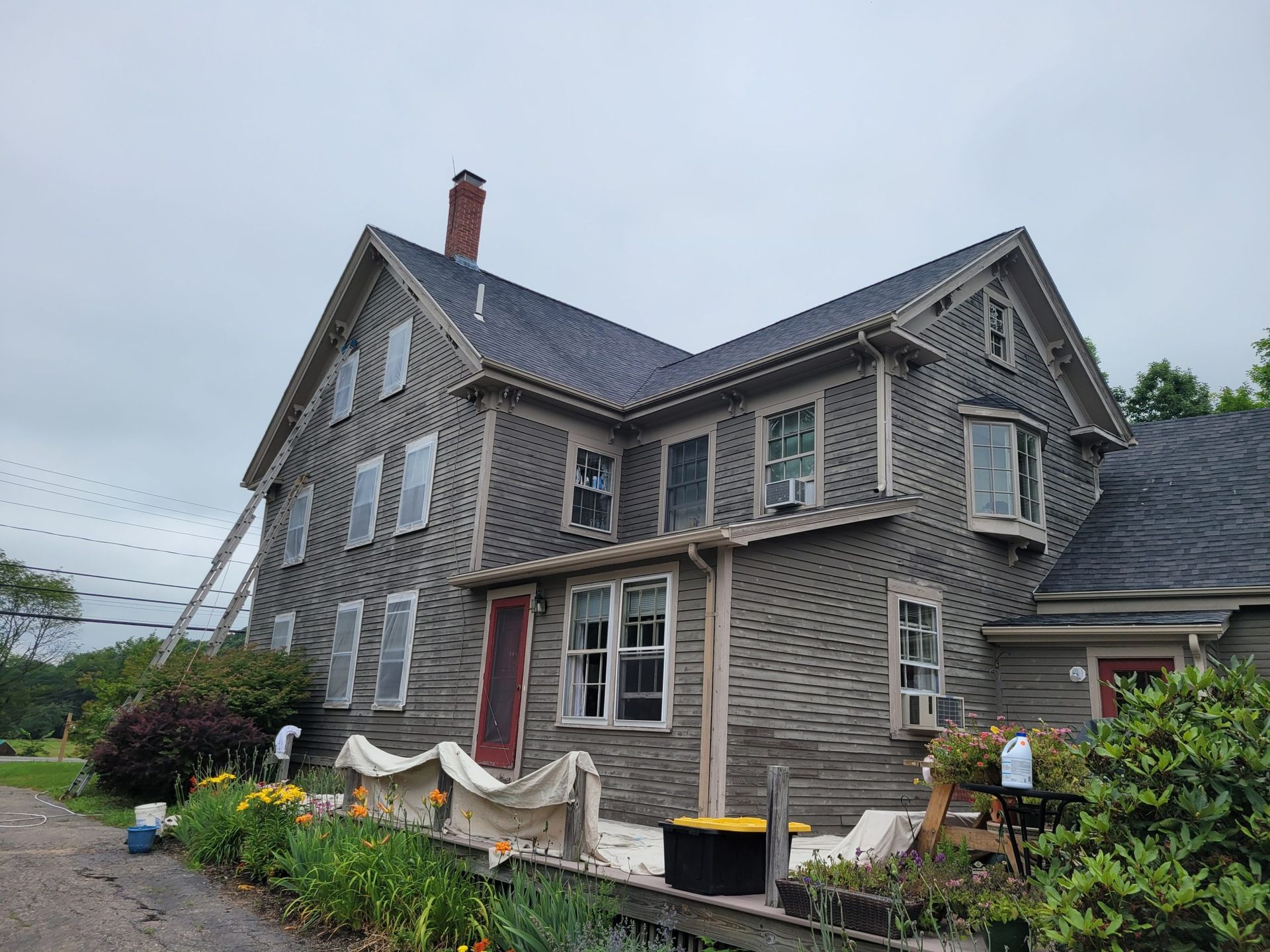 Weathered two-story house with gray siding; a worker on a ladder. Red door, gray roof, surrounded by plants.