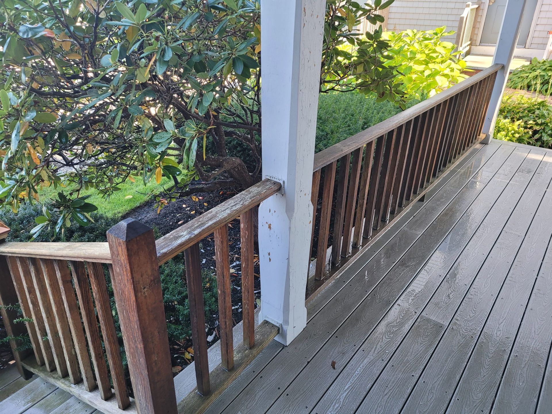 Wooden deck railing with weathered brown spindles. Gray deck boards, white post, and lush green plants.