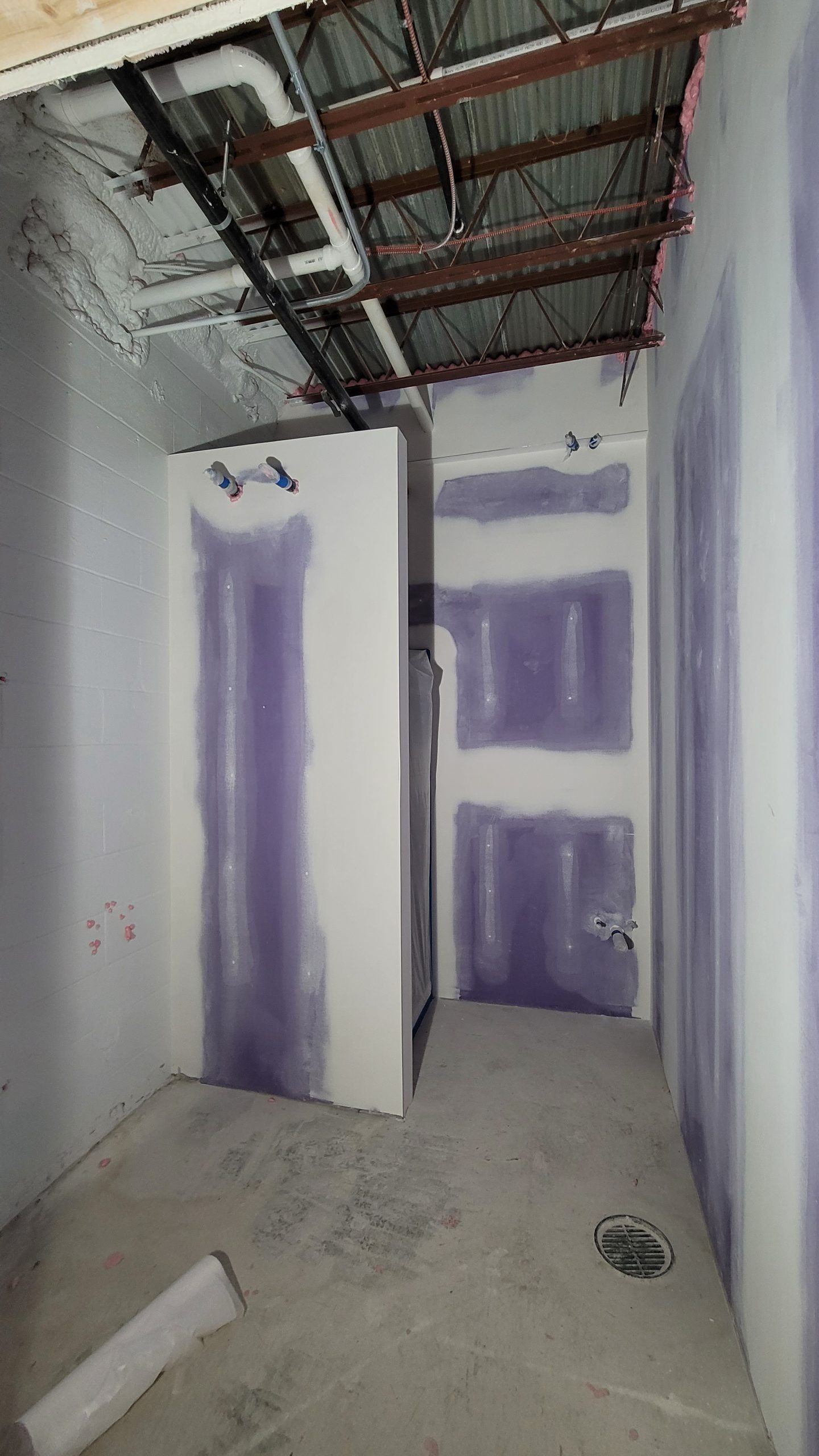 Interior construction of a small room. Drywall, purple joint compound, exposed ceiling beams and pipes.