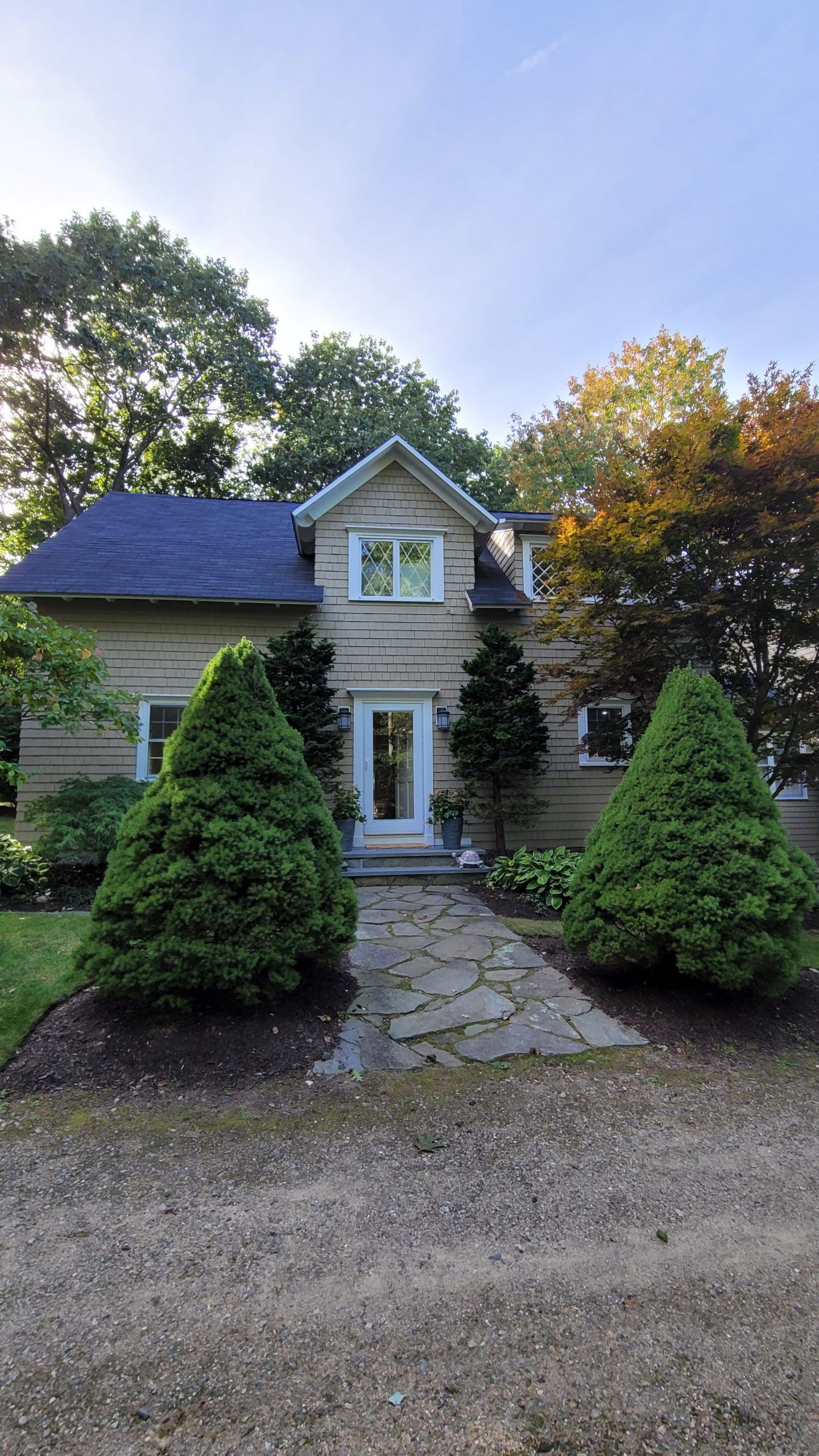 Beige house with a dark blue roof, stone path, two cone-shaped green bushes, and autumn trees.