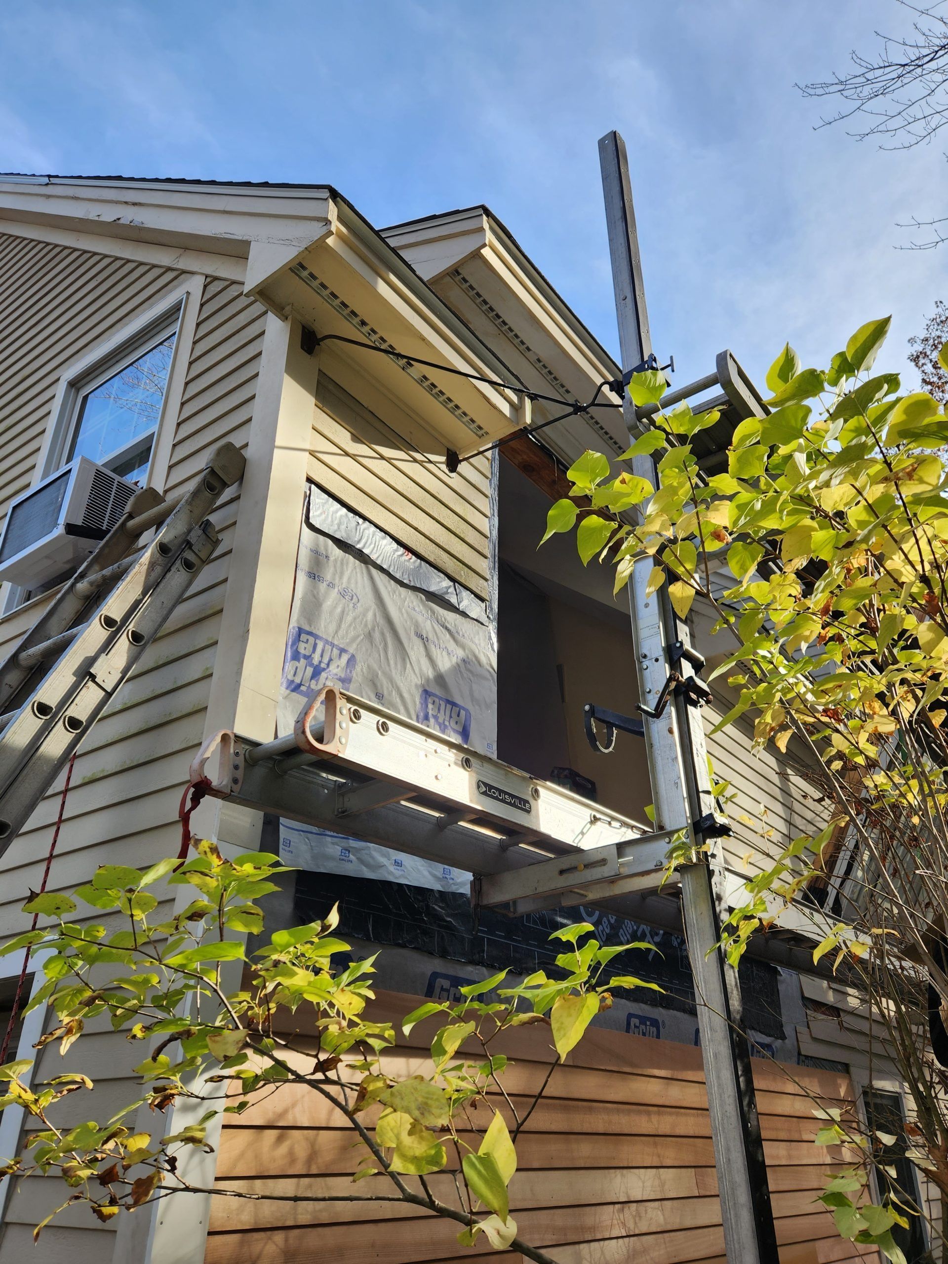 House exterior under construction, scaffolding, ladder, wood siding, clear sky.
