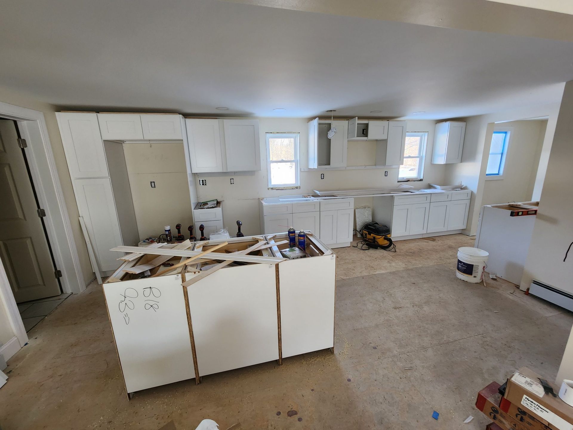 Kitchen under renovation with white cabinets, unfinished floors, and exposed walls.