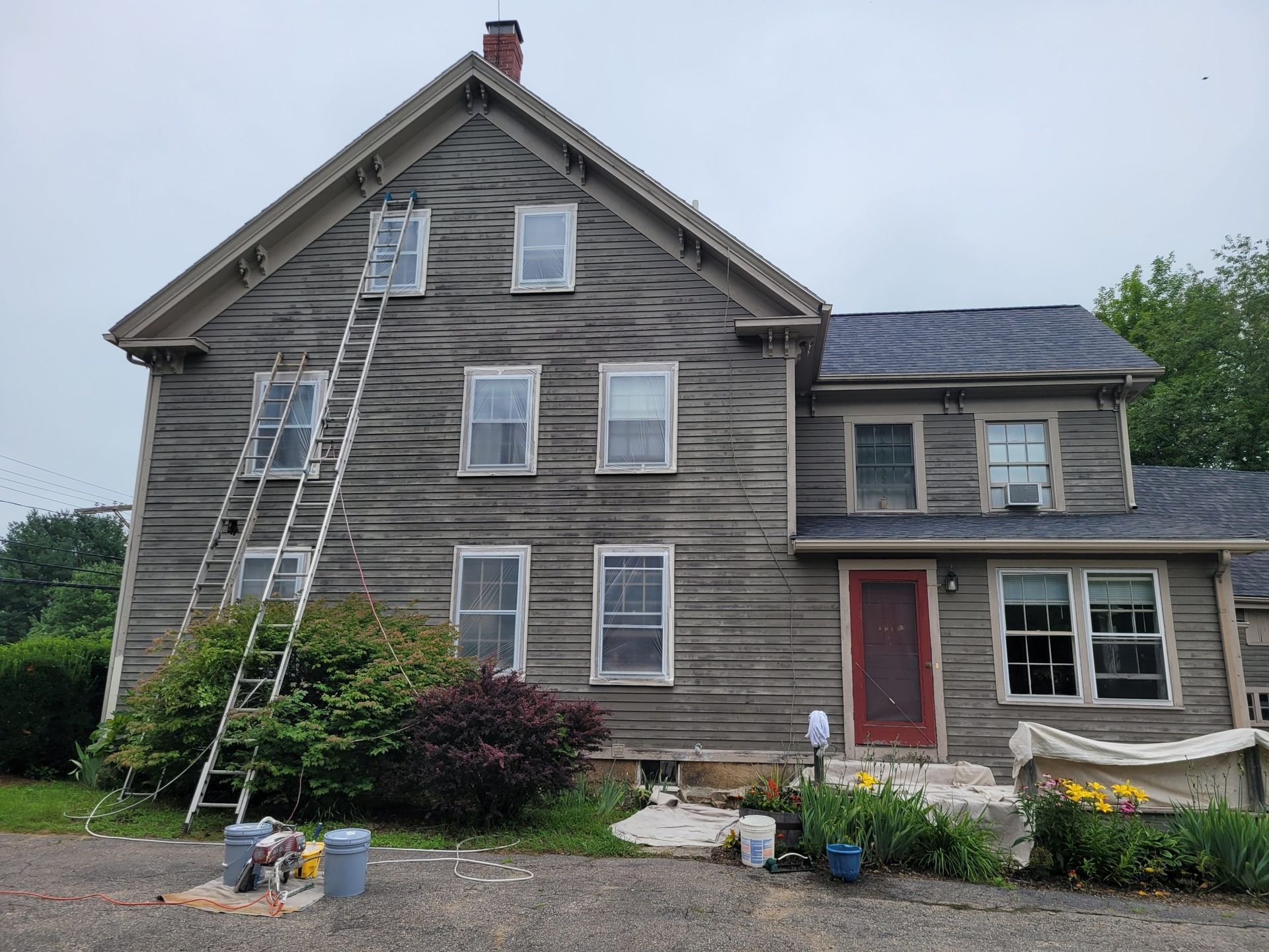 Two-story weathered gray house with ladder, undergoing exterior work. Red door, blue bucket, and flowers.
