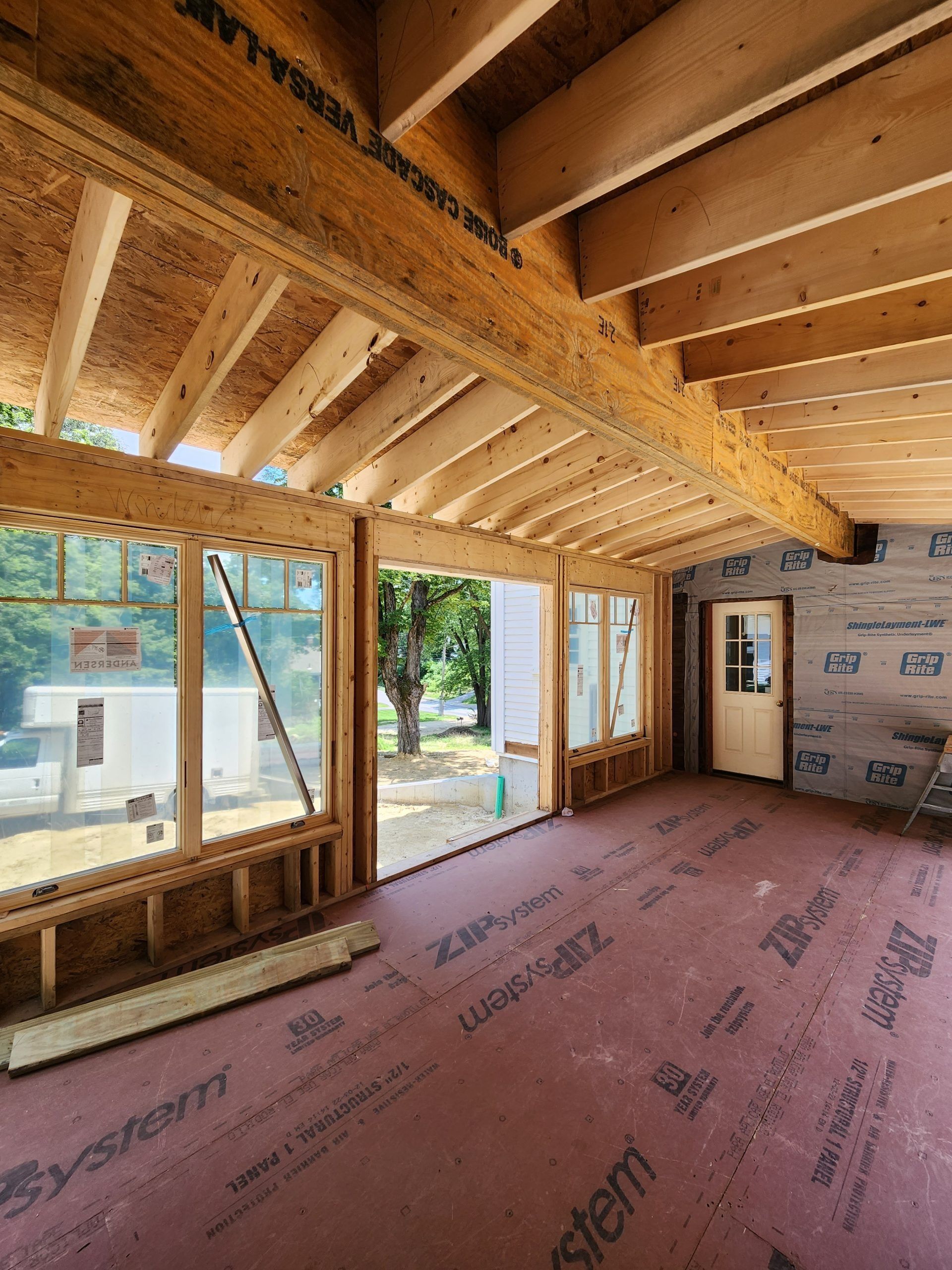 Interior of a room under construction, with wooden beams, large windows, and an unfinished floor.