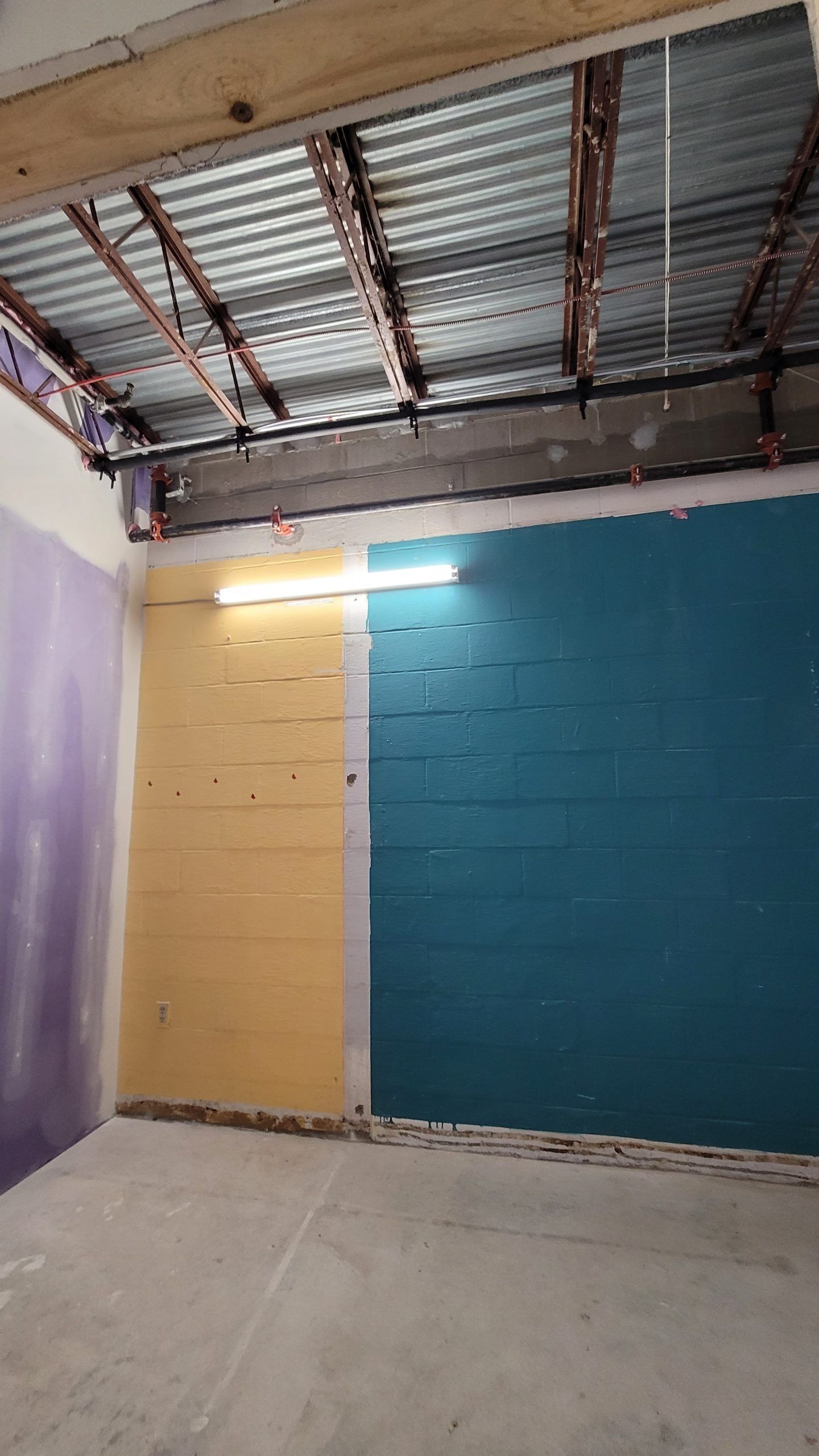 Room with unfinished ceiling and walls painted yellow, teal, and purple. A fluorescent light is on.