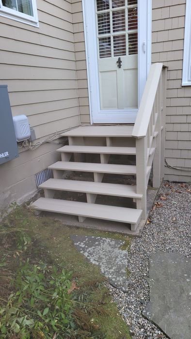 Beige wooden steps leading up to a white door with a glass pane, beside a beige clapboard house.