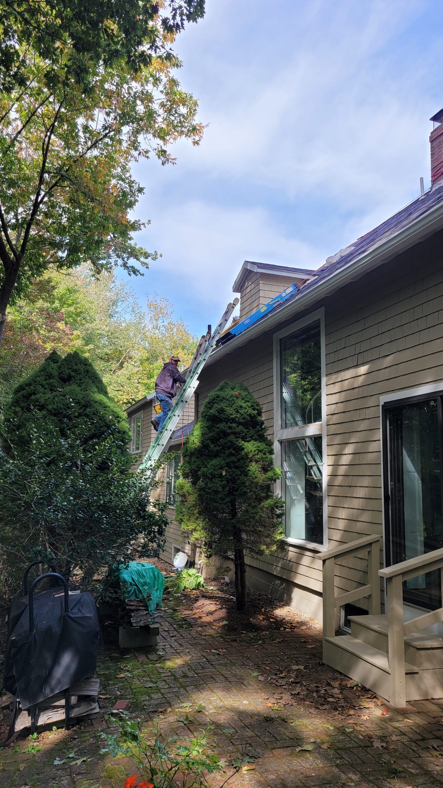 Person on a ladder, working on a roof. House exterior with trees and blue sky.